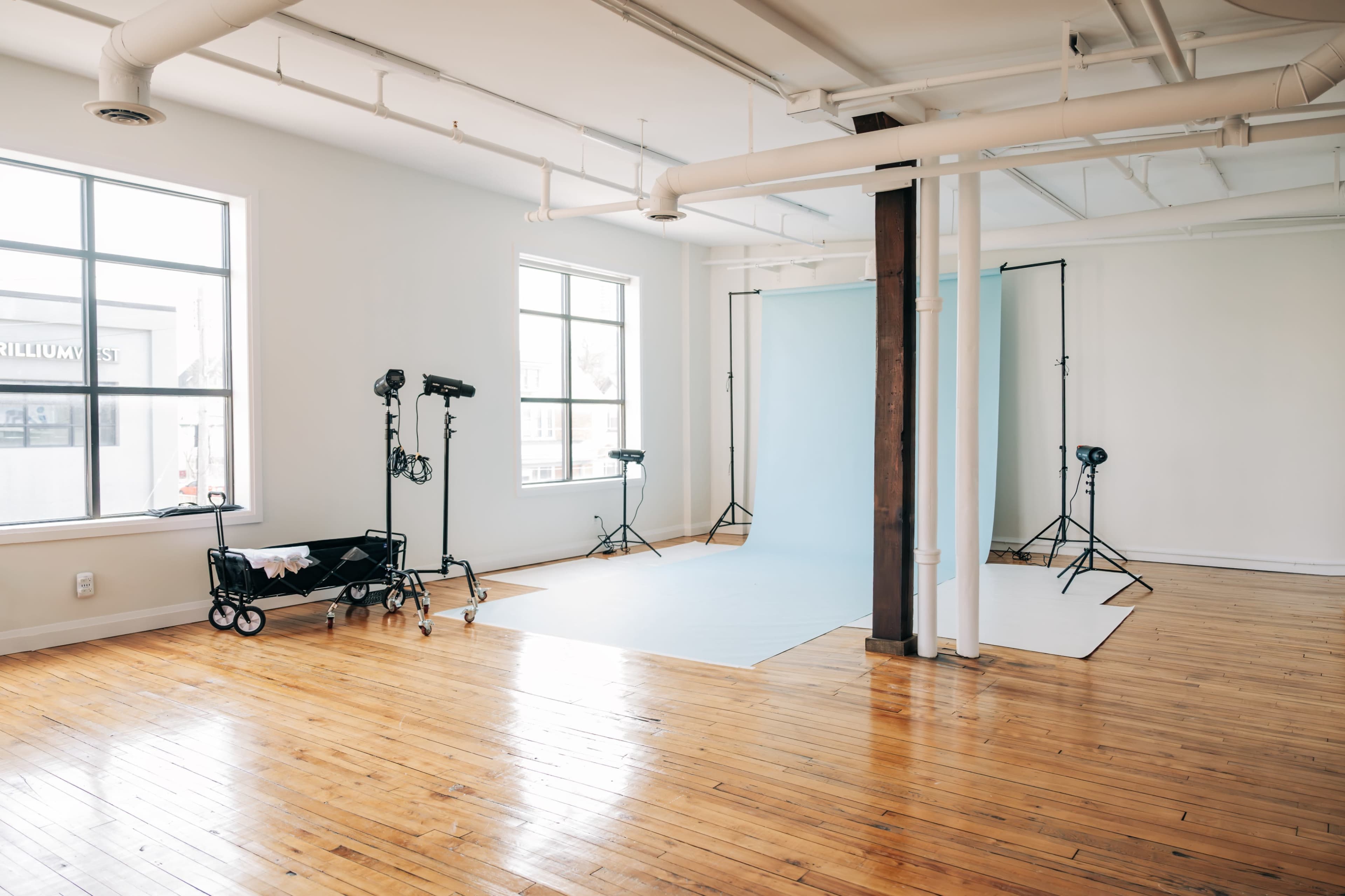 A spacious photography studio with large windows, wooden floors, and backdrops set up on stands.