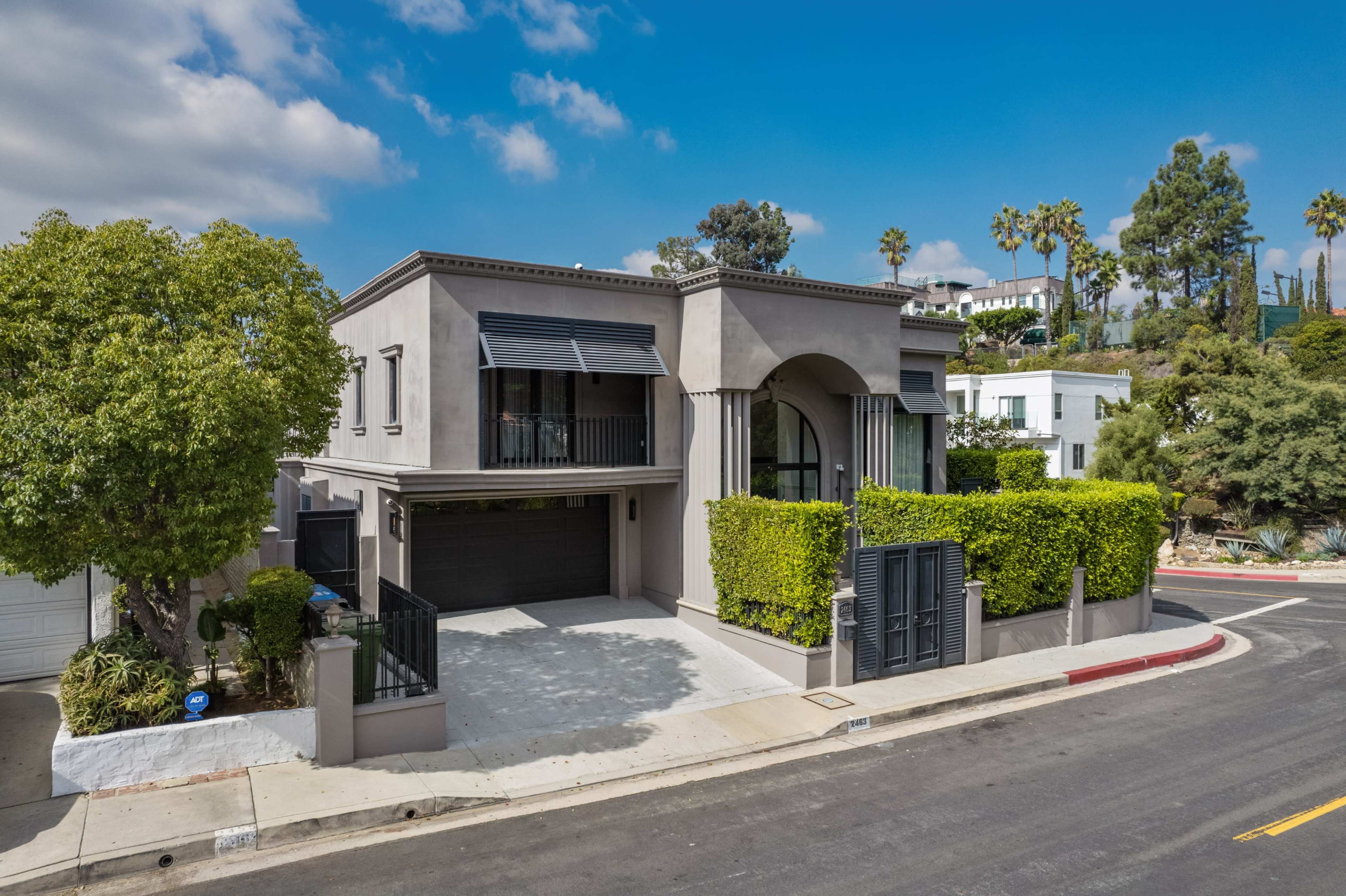 A modern, two-story house with a gray exterior and lush greenery surrounding the entrance sits at the corner of a quiet street.