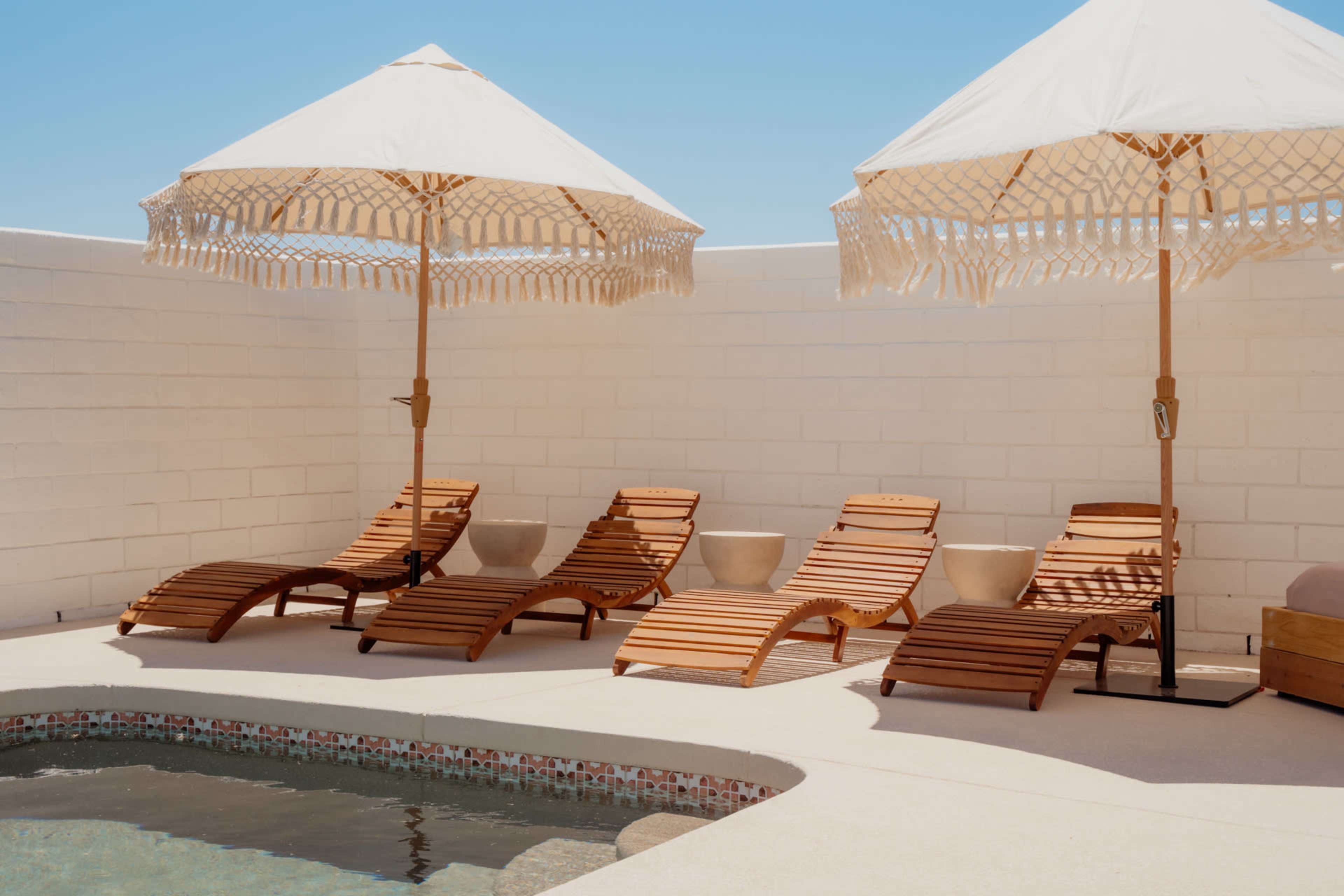 Four wooden lounge chairs with white umbrellas are arranged beside a small swimming pool in a sunlit area enclosed by a white wall.