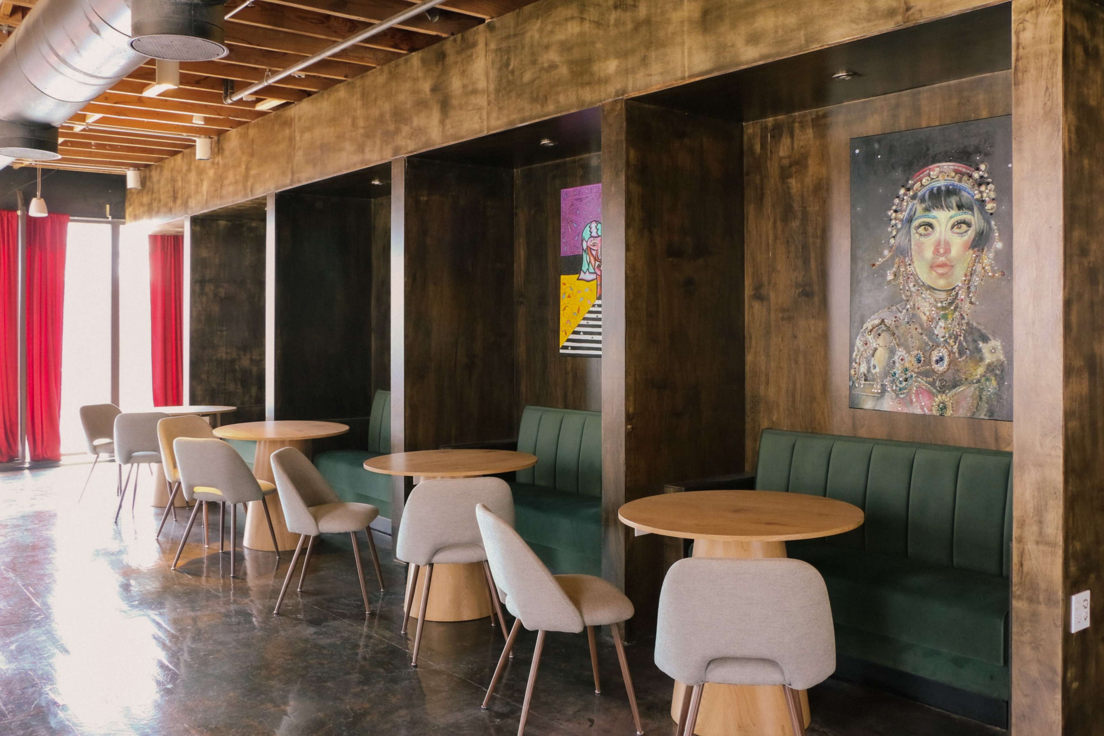 The image shows a modern dining area featuring wooden booths with green upholstery, round tables, and colorful artwork on the walls, alongside red curtains and exposed ceiling beams.