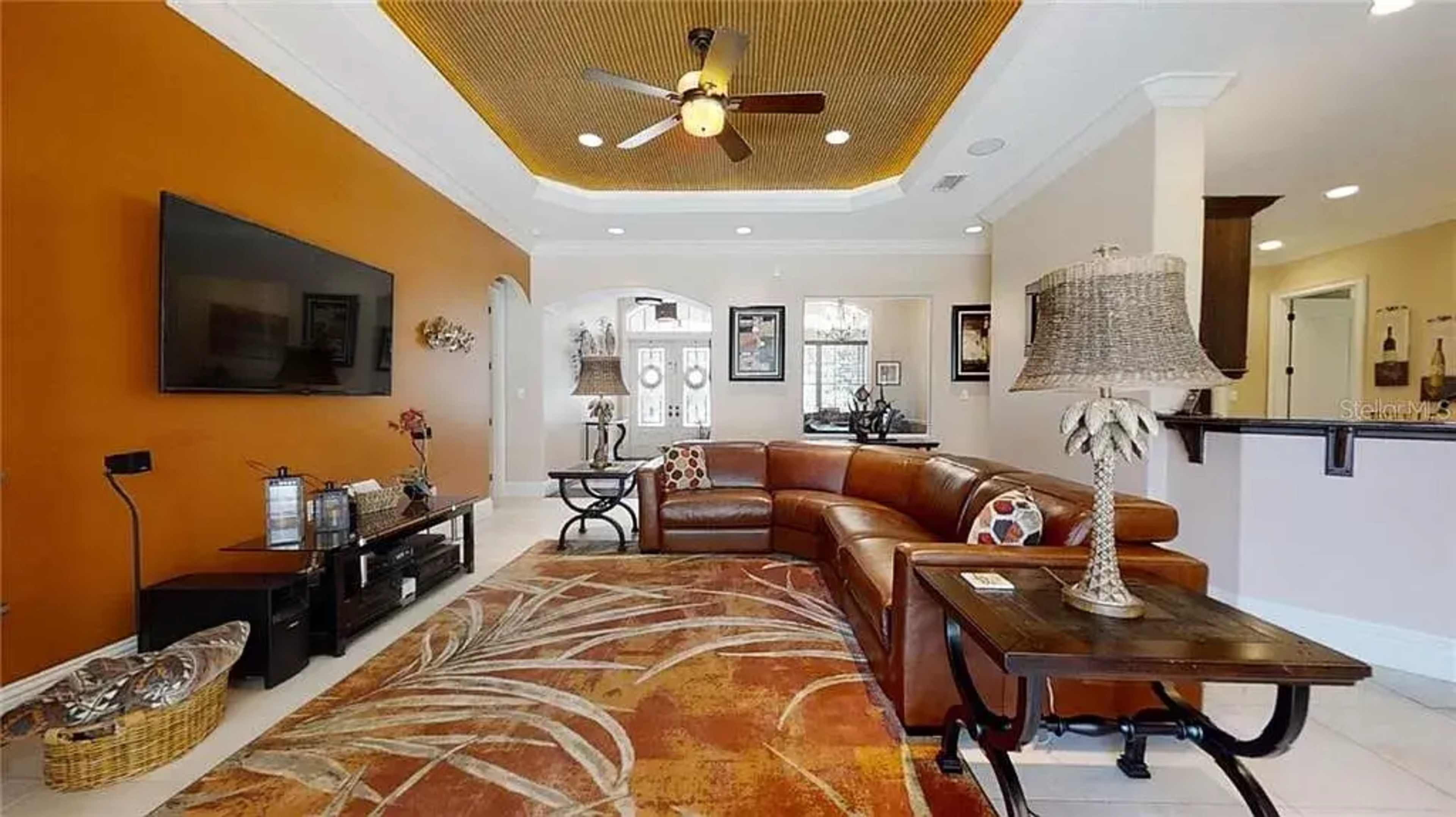 The living room features a brown sectional sofa arranged around a patterned area rug, with an orange accent wall and a ceiling fan above.