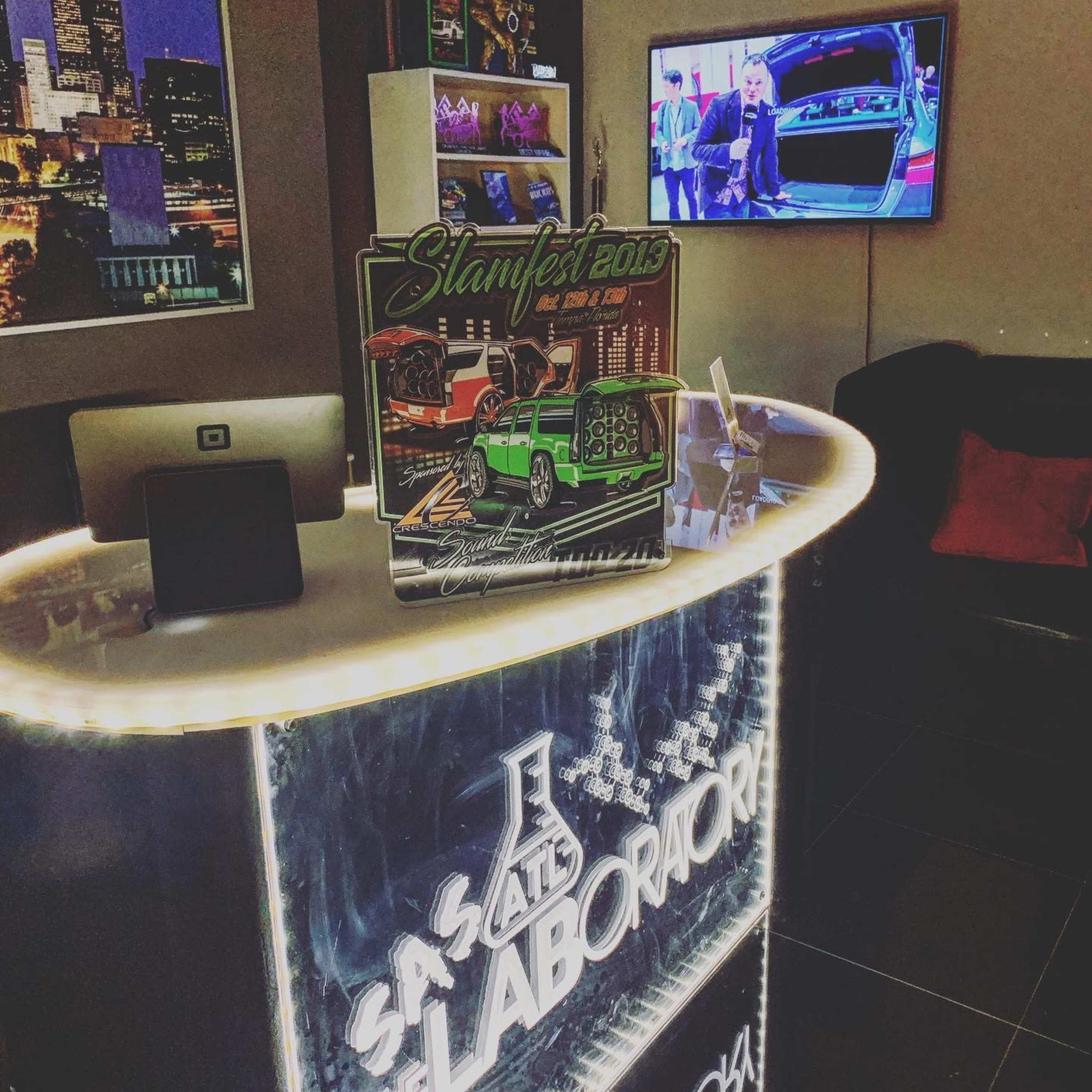 A brightly lit reception desk in a modern office setting, featuring a promotional poster for "Slamfest 2013" and a television displaying a vehicle-related program.
