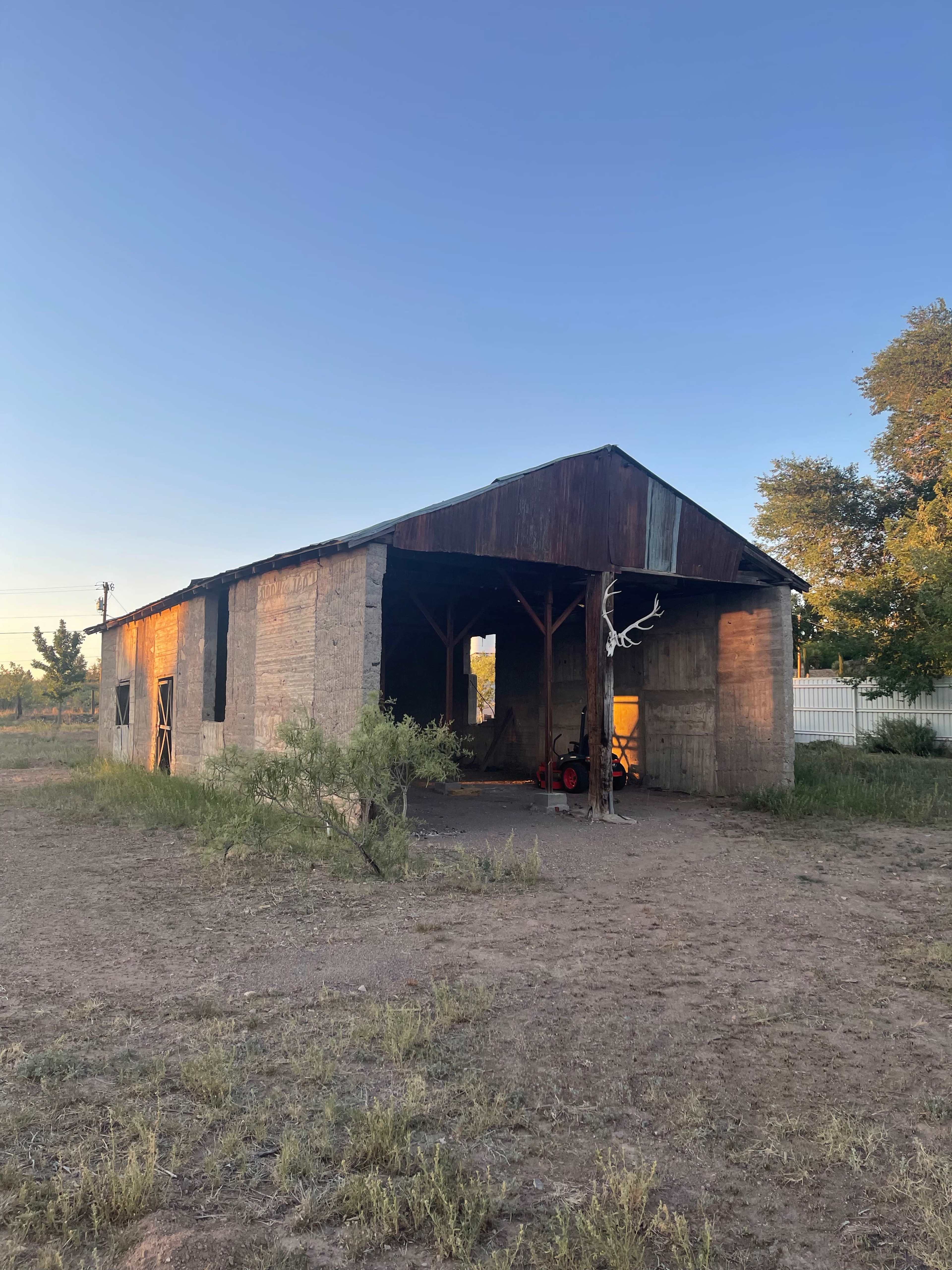 The image shows a weathered barn with a rusty roof and an open side, set in a barren landscape with sparse vegetation under a clear blue sky.