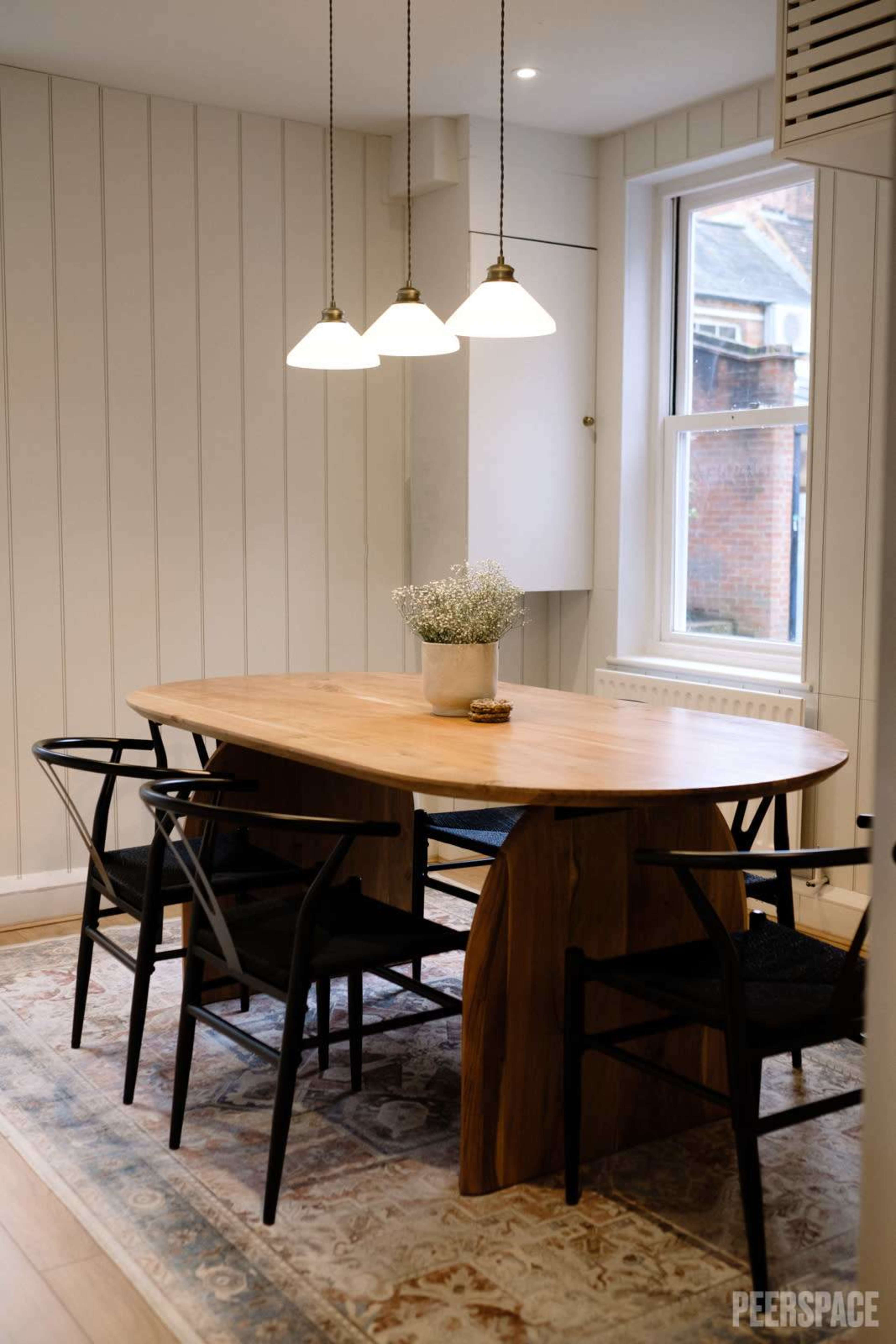 A light wood dining table with black chairs is centered under three pendant lights in a room with white paneled walls and a window.