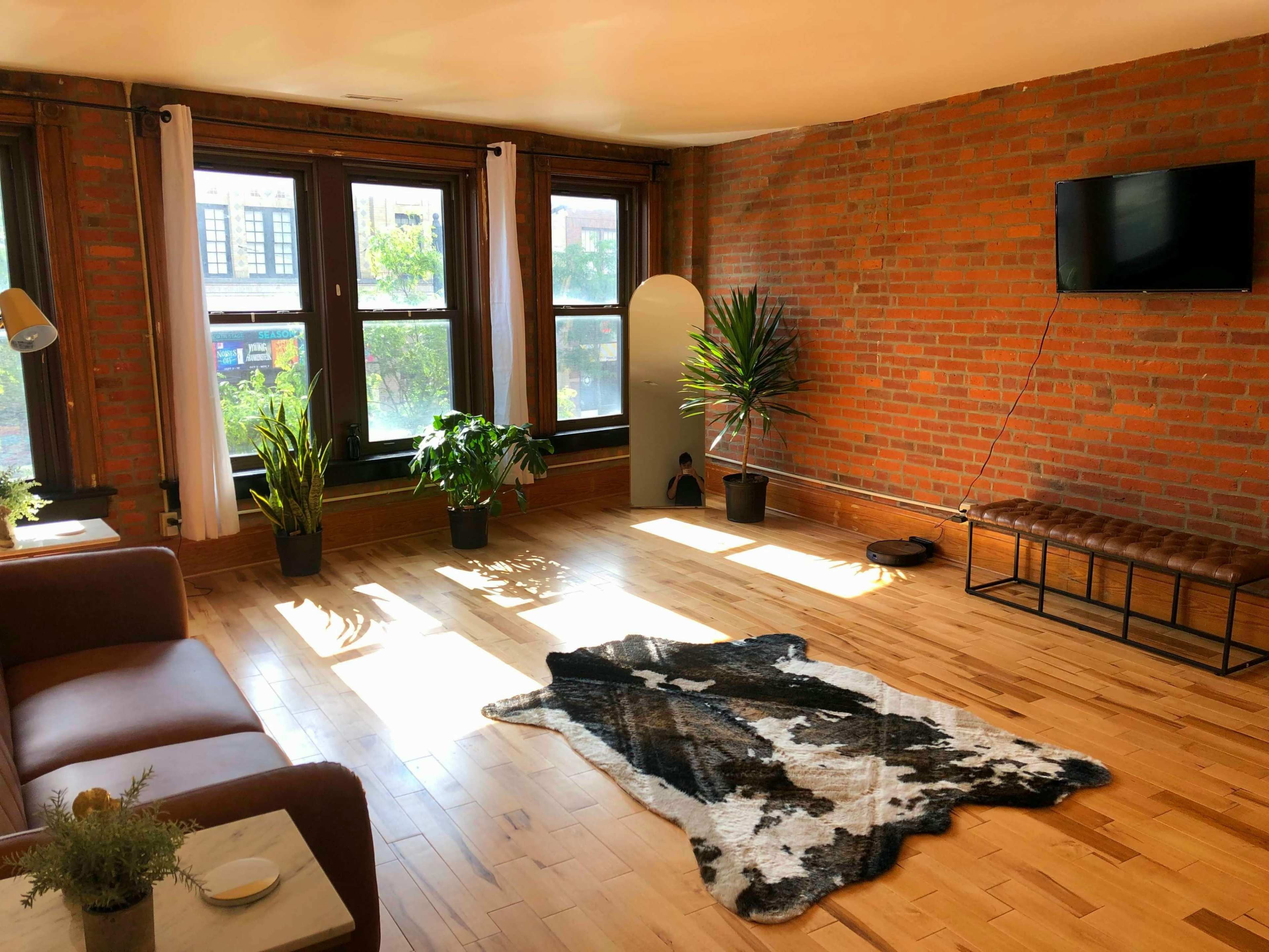 A sunlit room features hardwood floors, a brown sofa, indoor plants, and a TV mounted on a brick wall.