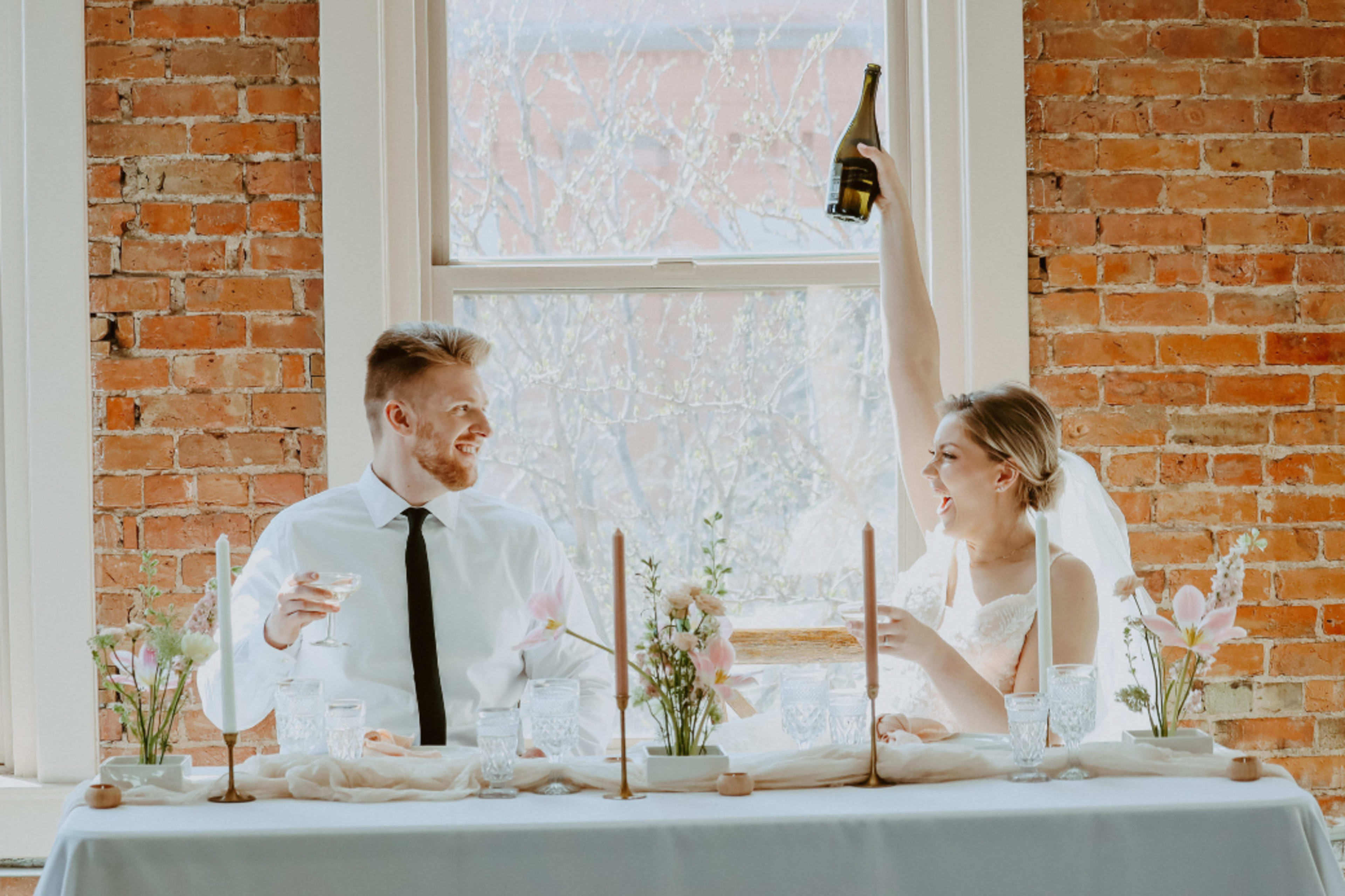 A bride and groom celebrate at a table adorned with flowers and candles, with the bride joyfully raising a champagne bottle.