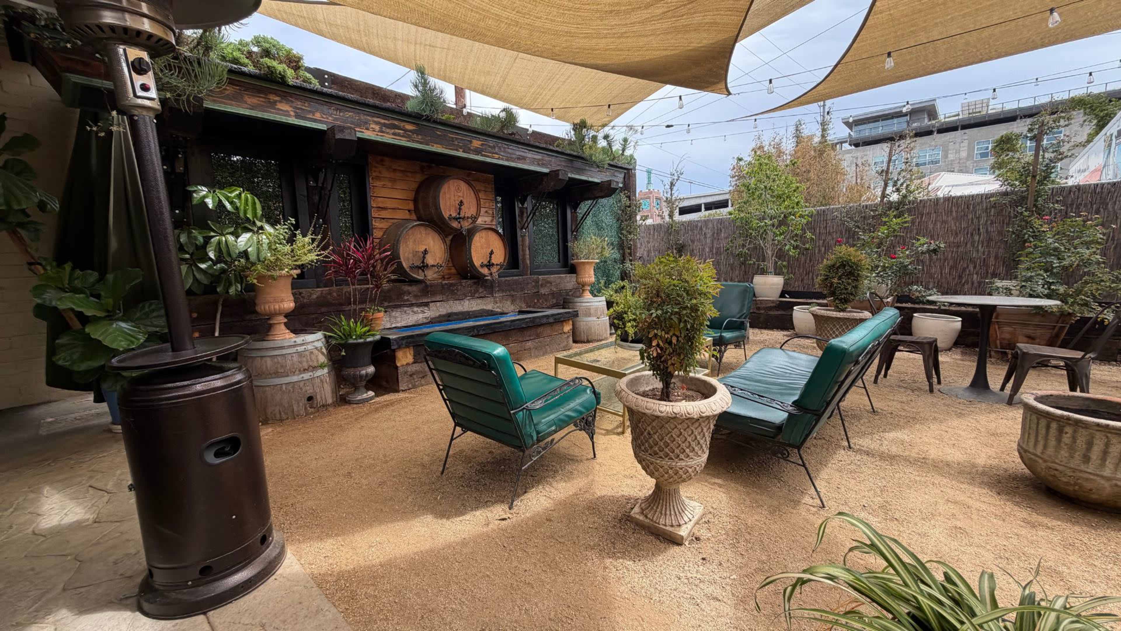 The image shows a cozy outdoor seating area with green chairs and a table, surrounded by plants and decorative barrels.