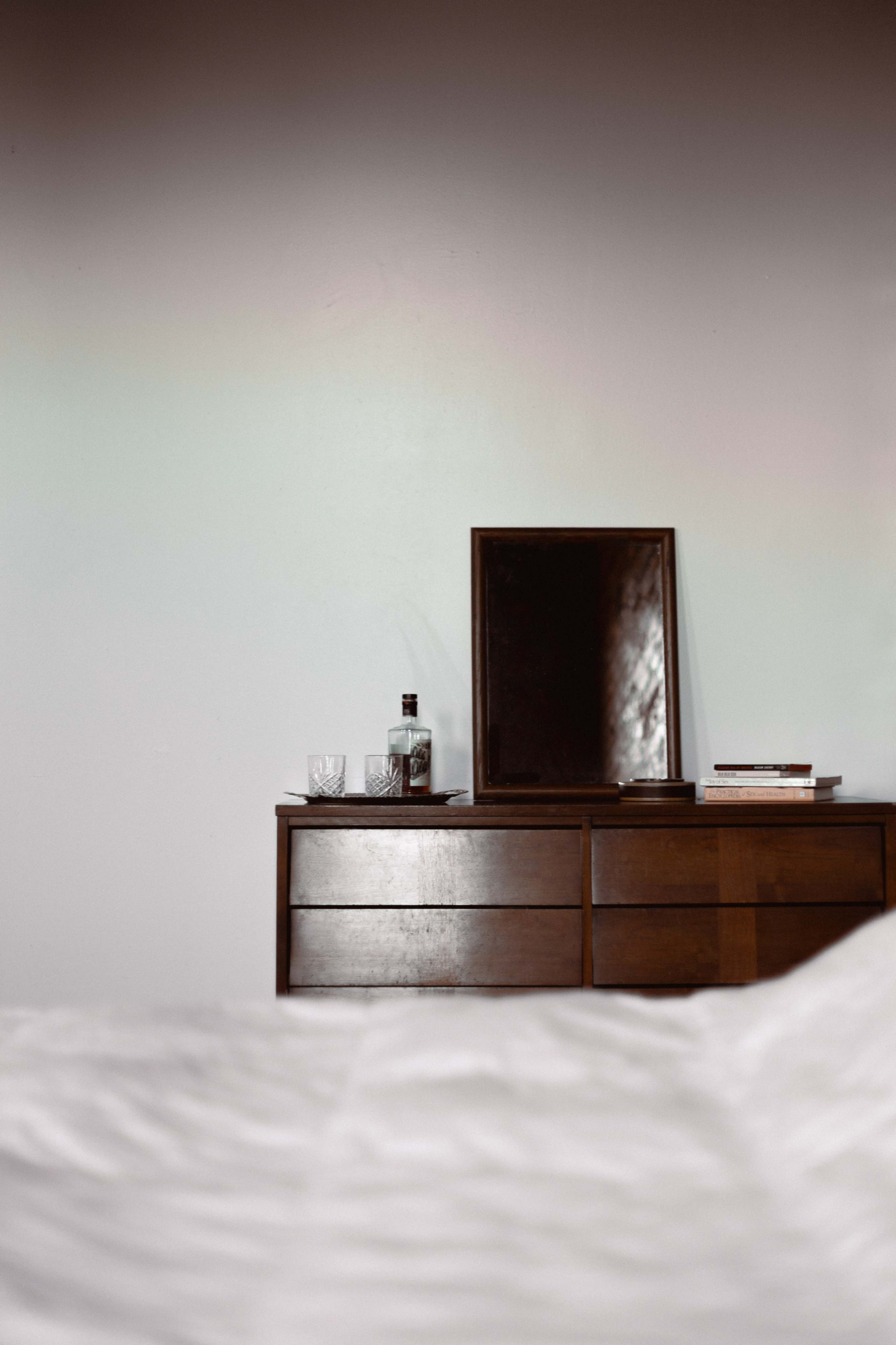 A wooden dresser with a mirror sits against a wall, accompanied by a bottle and glassware on top.