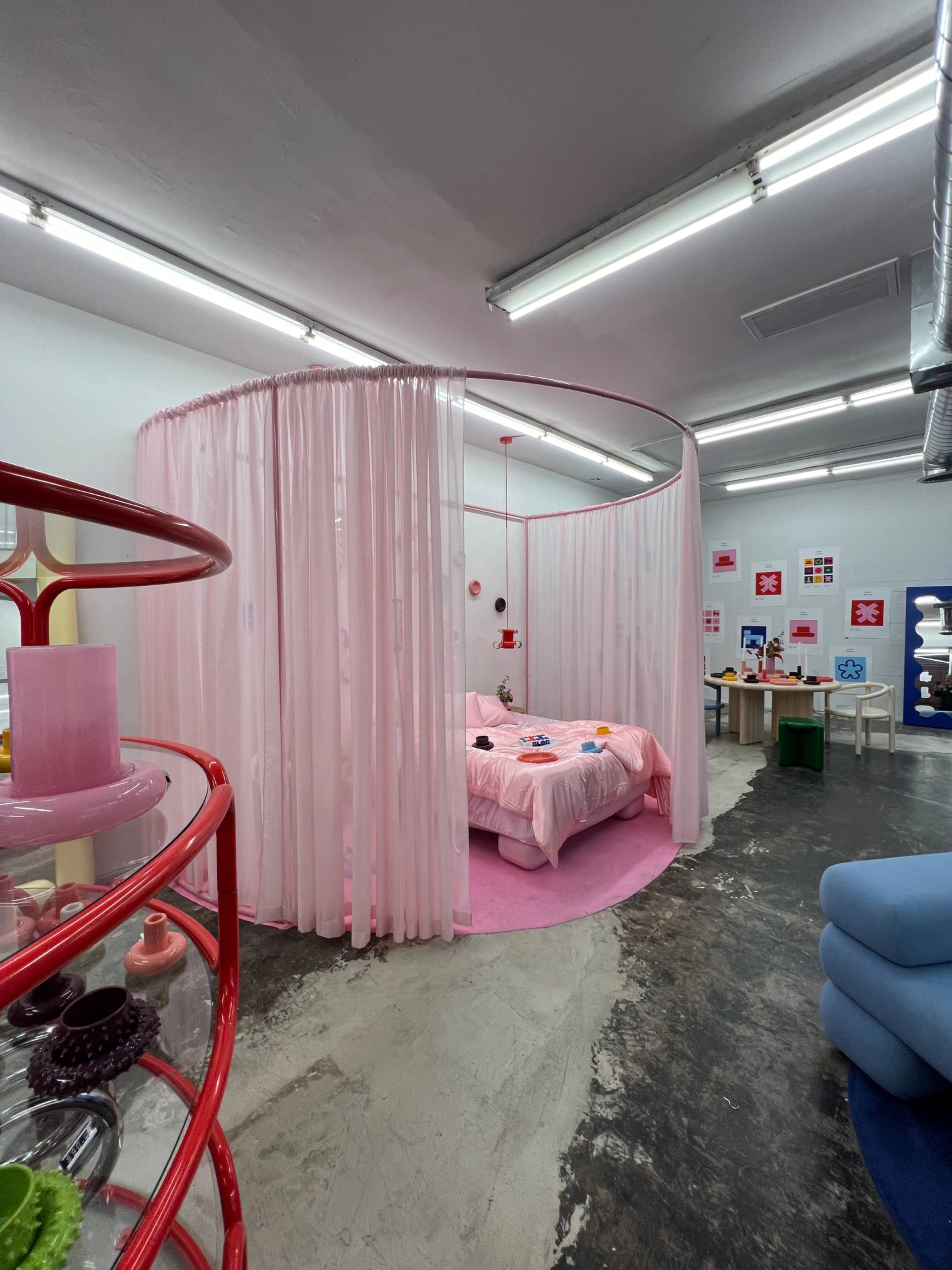 A room with a pink bed surrounded by sheer curtains, located in a space with bright fluorescent lighting and minimal furniture.