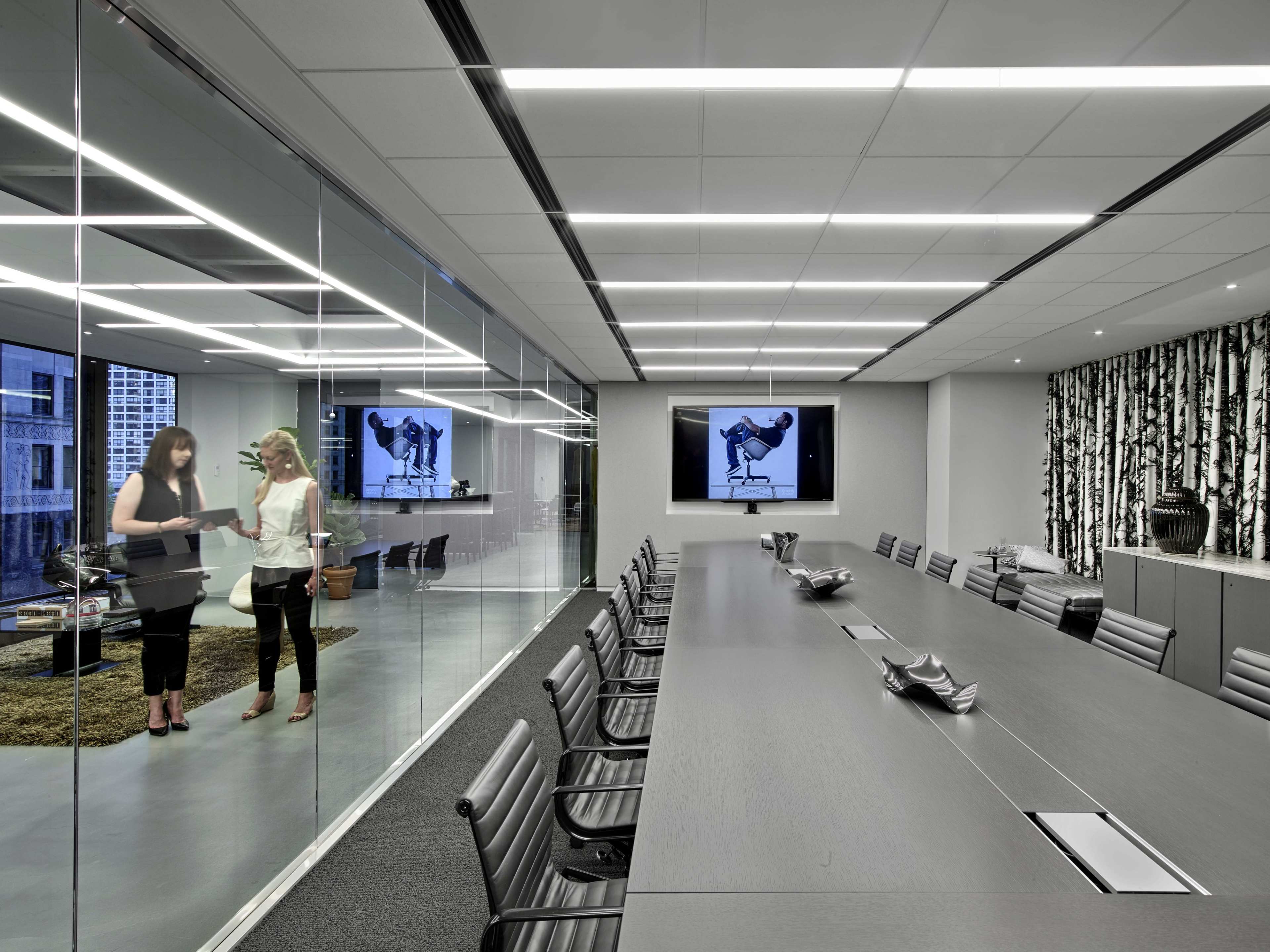 A modern conference room features a long table, glass walls, and two women discussing beside a large screen displaying a black-and-white image.
