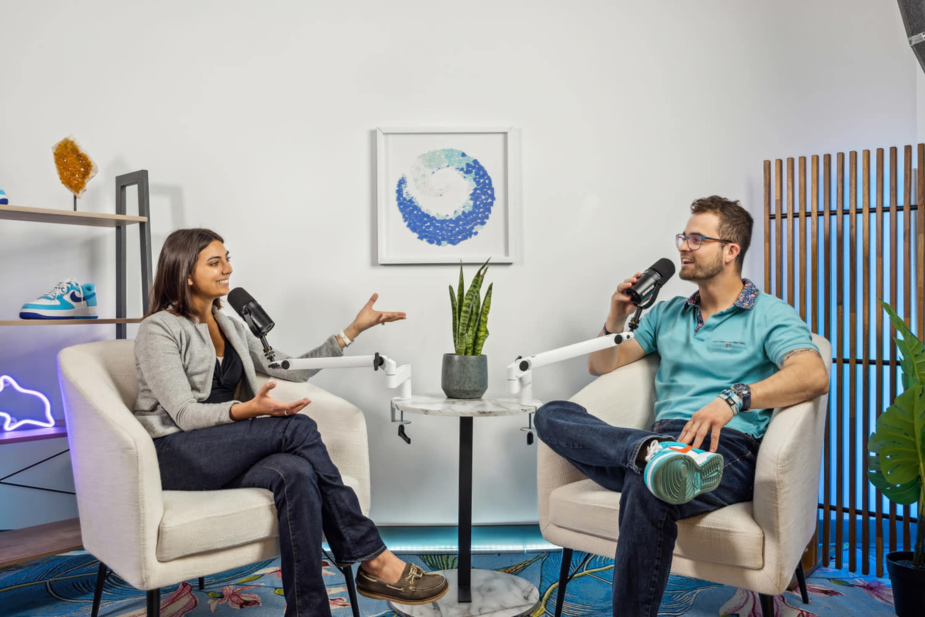 Two individuals are seated in modern chairs with microphones, engaged in a conversation in a well-decorated studio space.