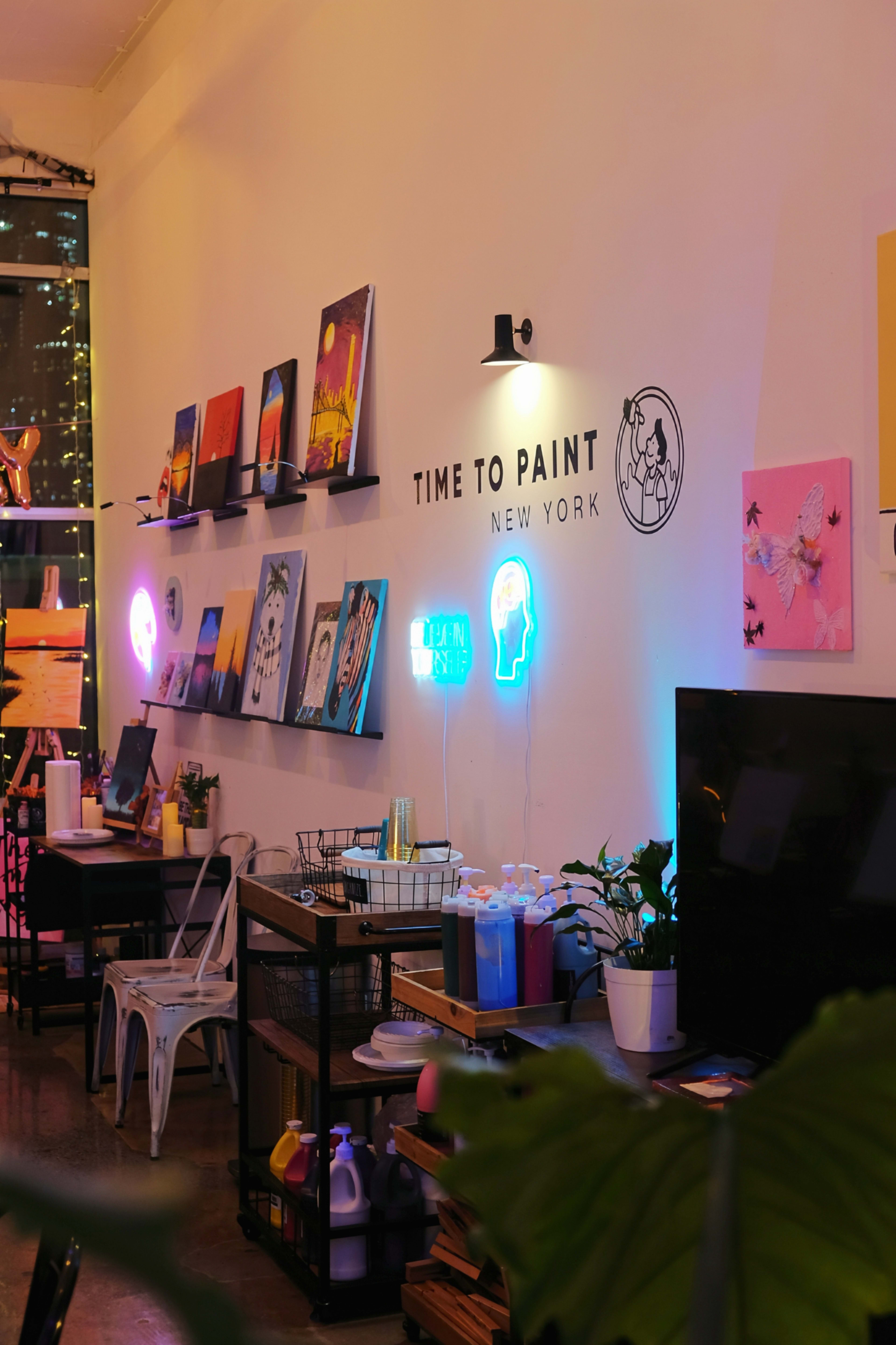 The image shows a cozy art studio in New York with brightly colored paintings displayed on the wall, neon signs, and creative supplies organized on shelves and tables.