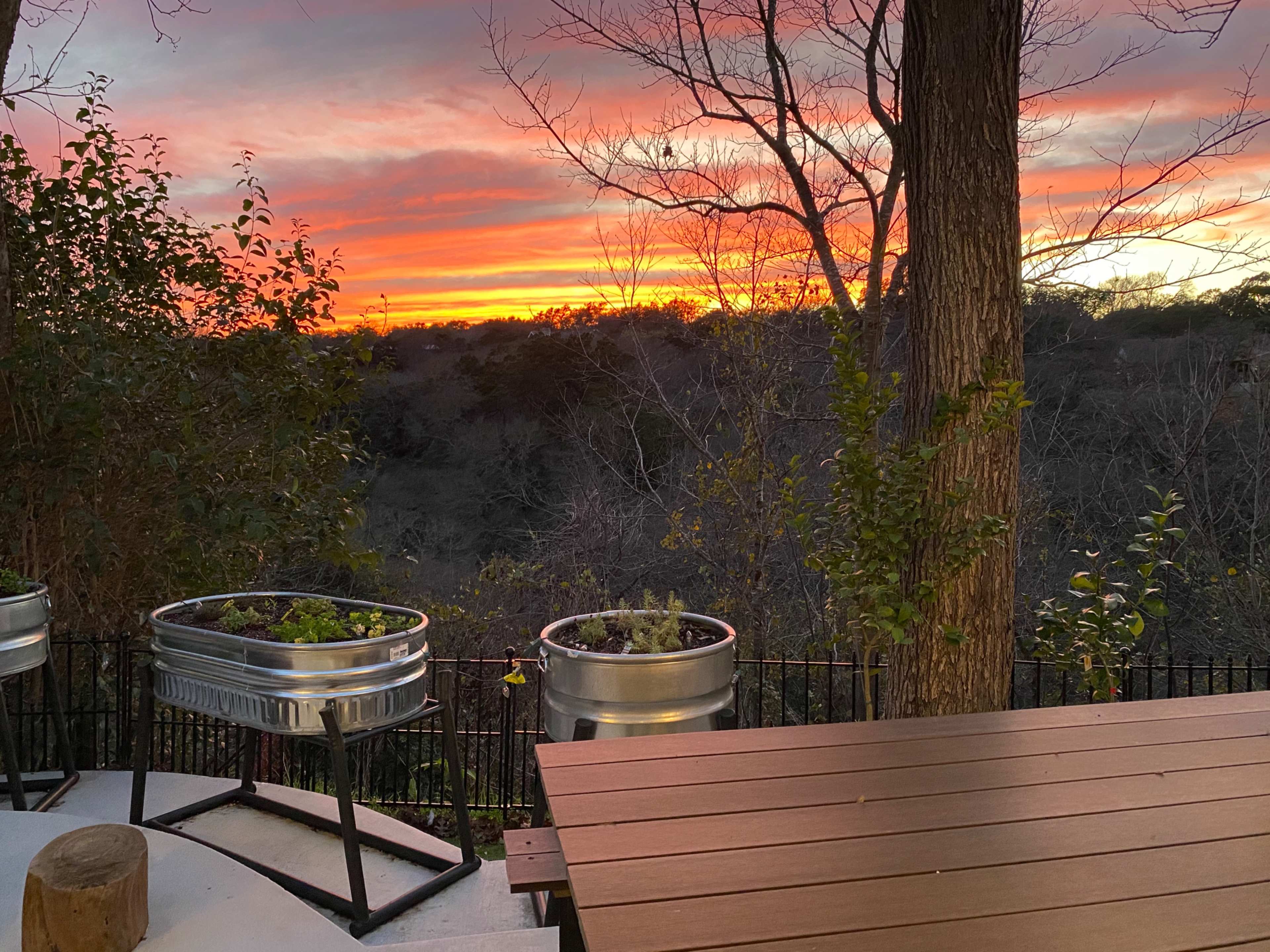The image shows a sunset over a landscape, with metal planters and a wooden table in the foreground.