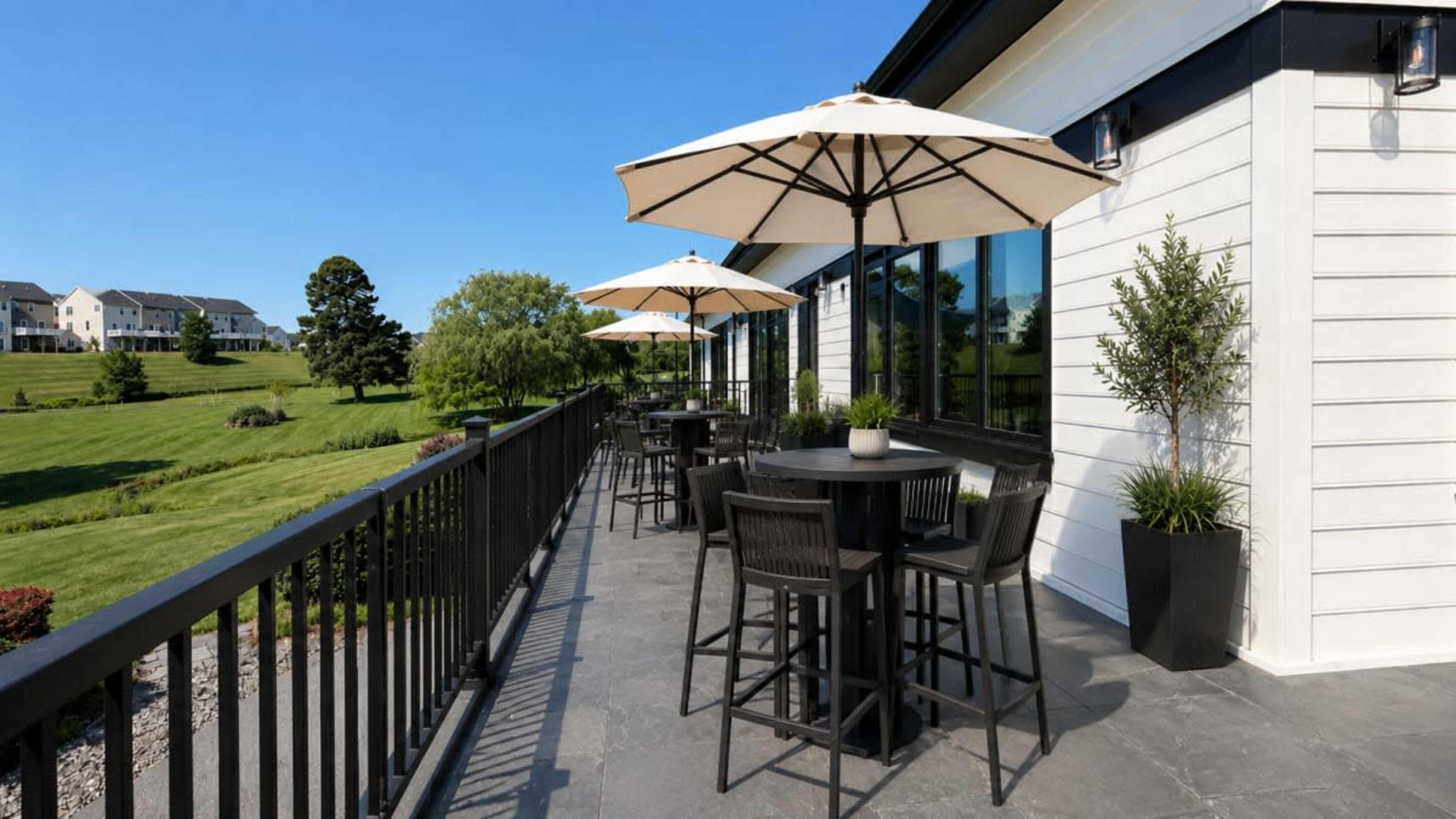 The image shows a patio area with tall black tables and chairs under large umbrellas, overlooking a green landscape and blue sky.