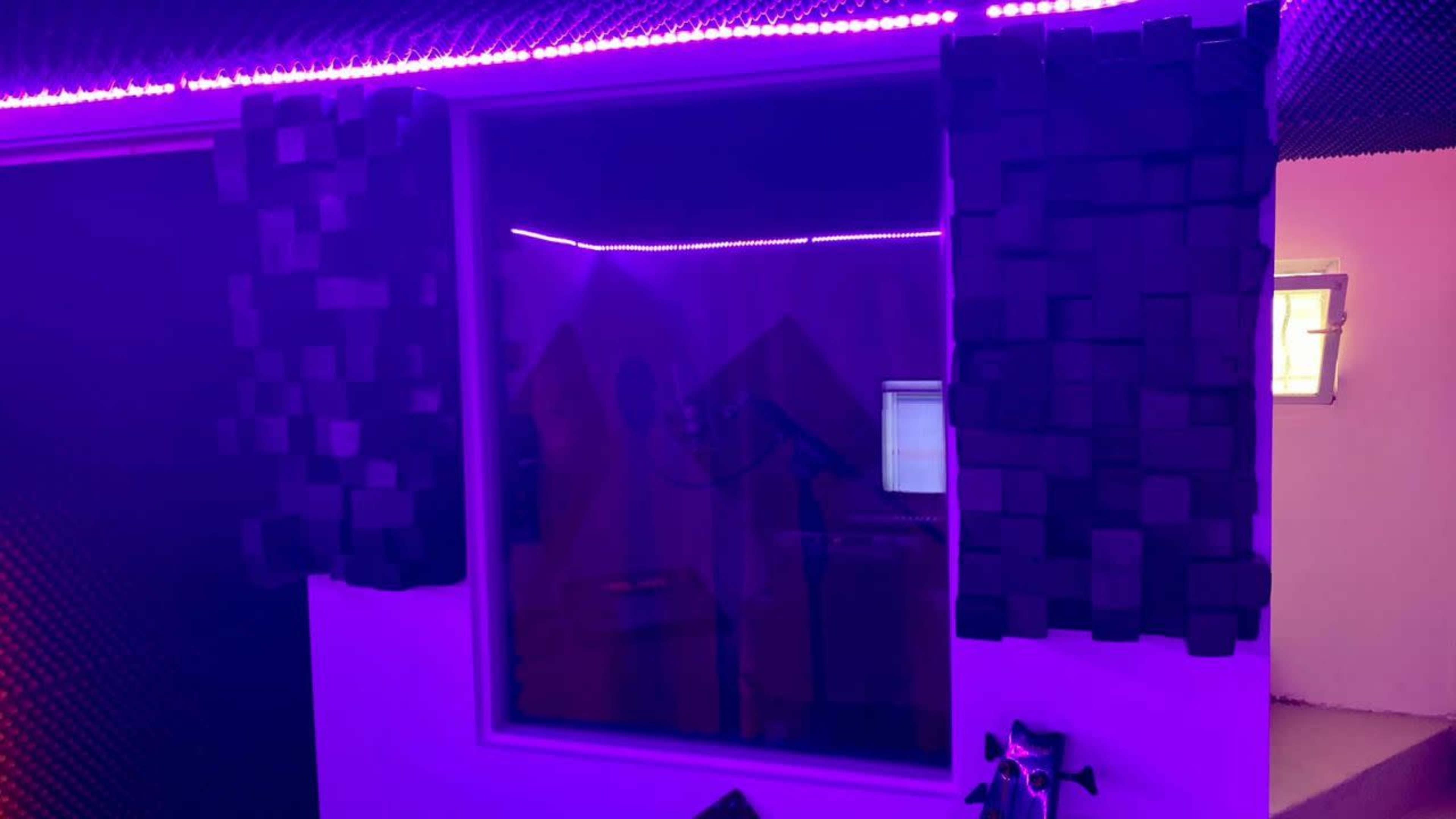 A soundproof recording studio with purple ambient lighting, featuring acoustic panels around a glass window and a microphone setup inside.