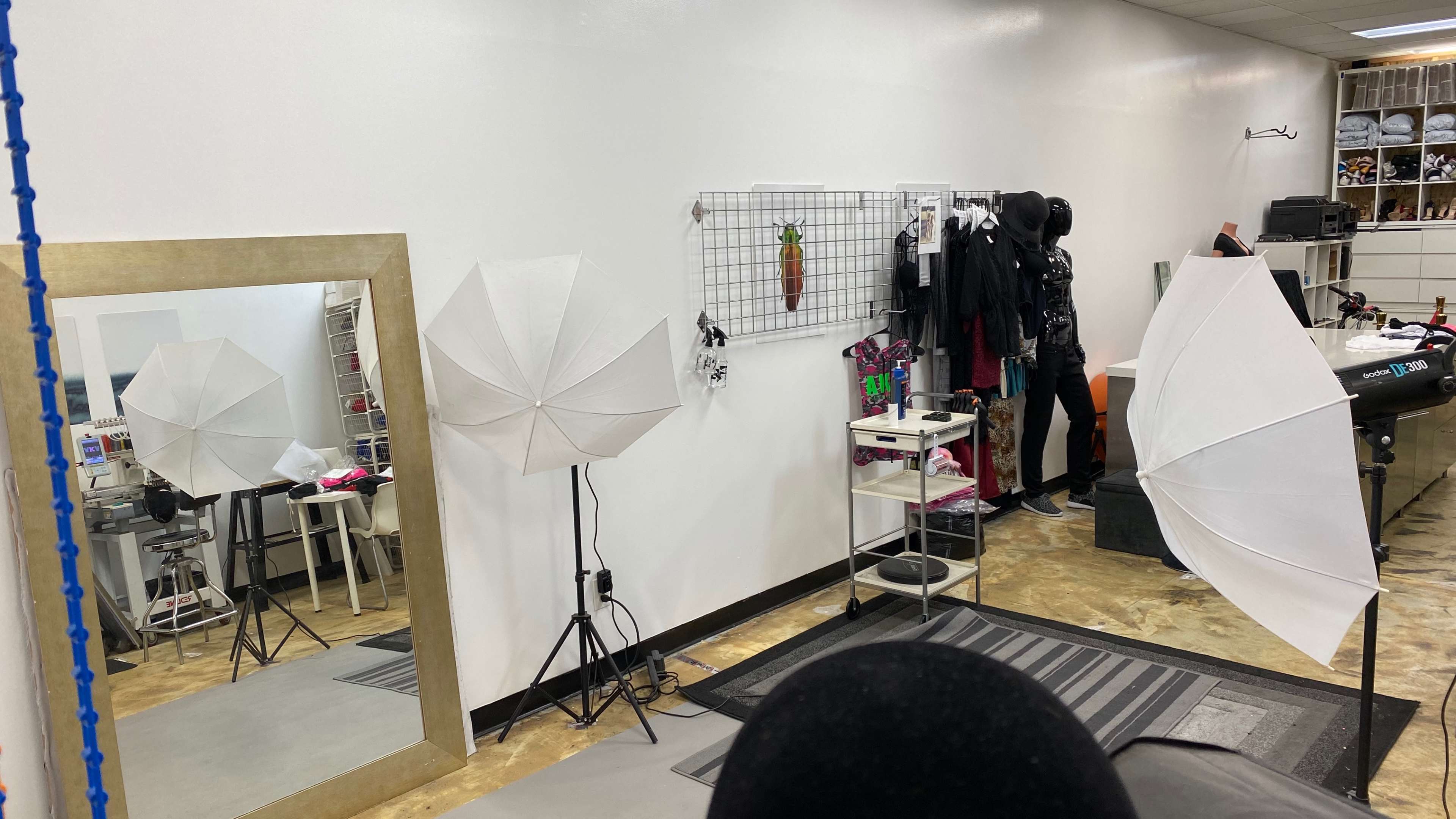 A photography studio setup with two large softboxes, a mirror, and various clothing items displayed on racks against a white wall.