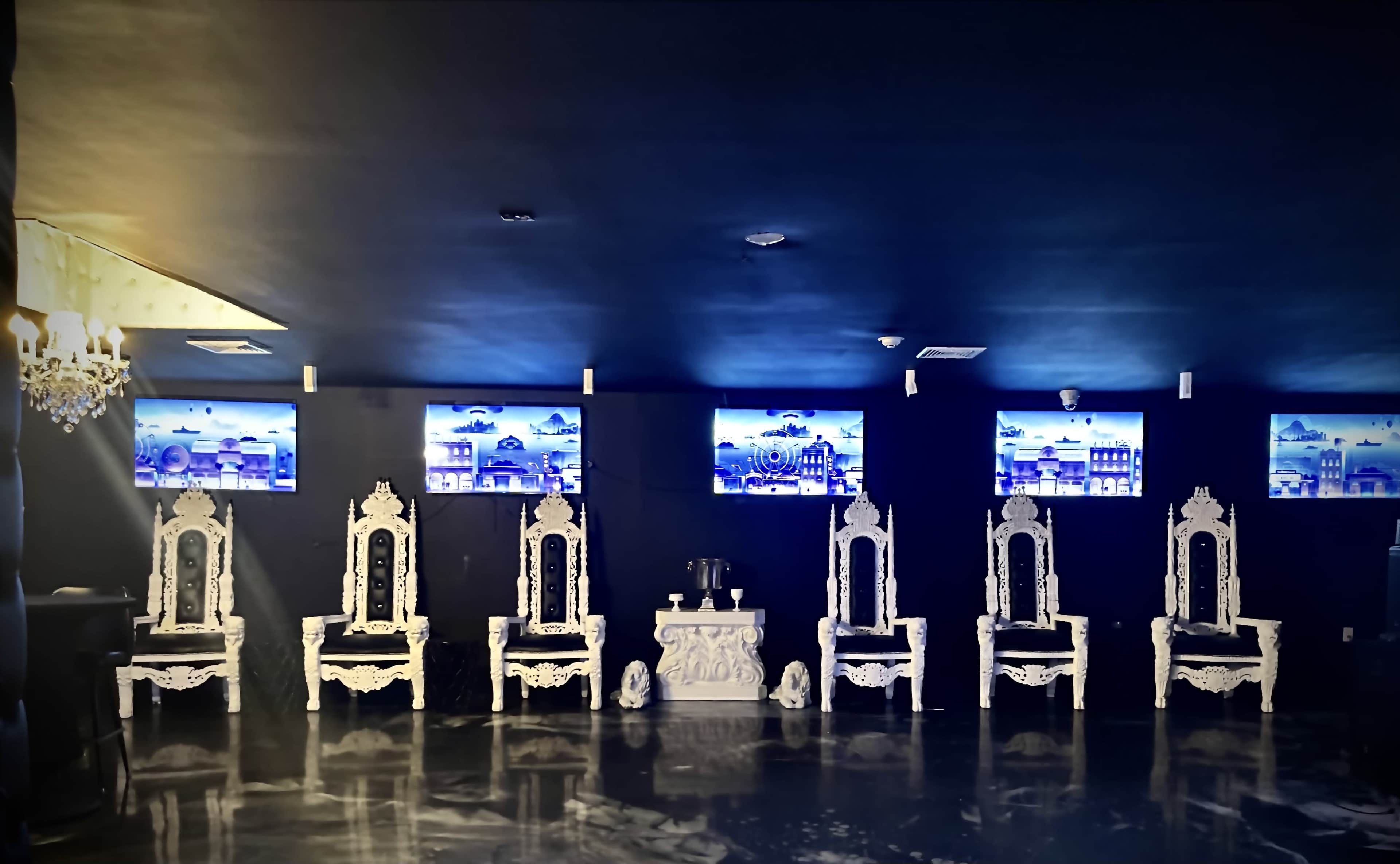 A dimly lit room with six ornate white chairs arranged around a central table, and three screens displaying images mounted on the dark walls.