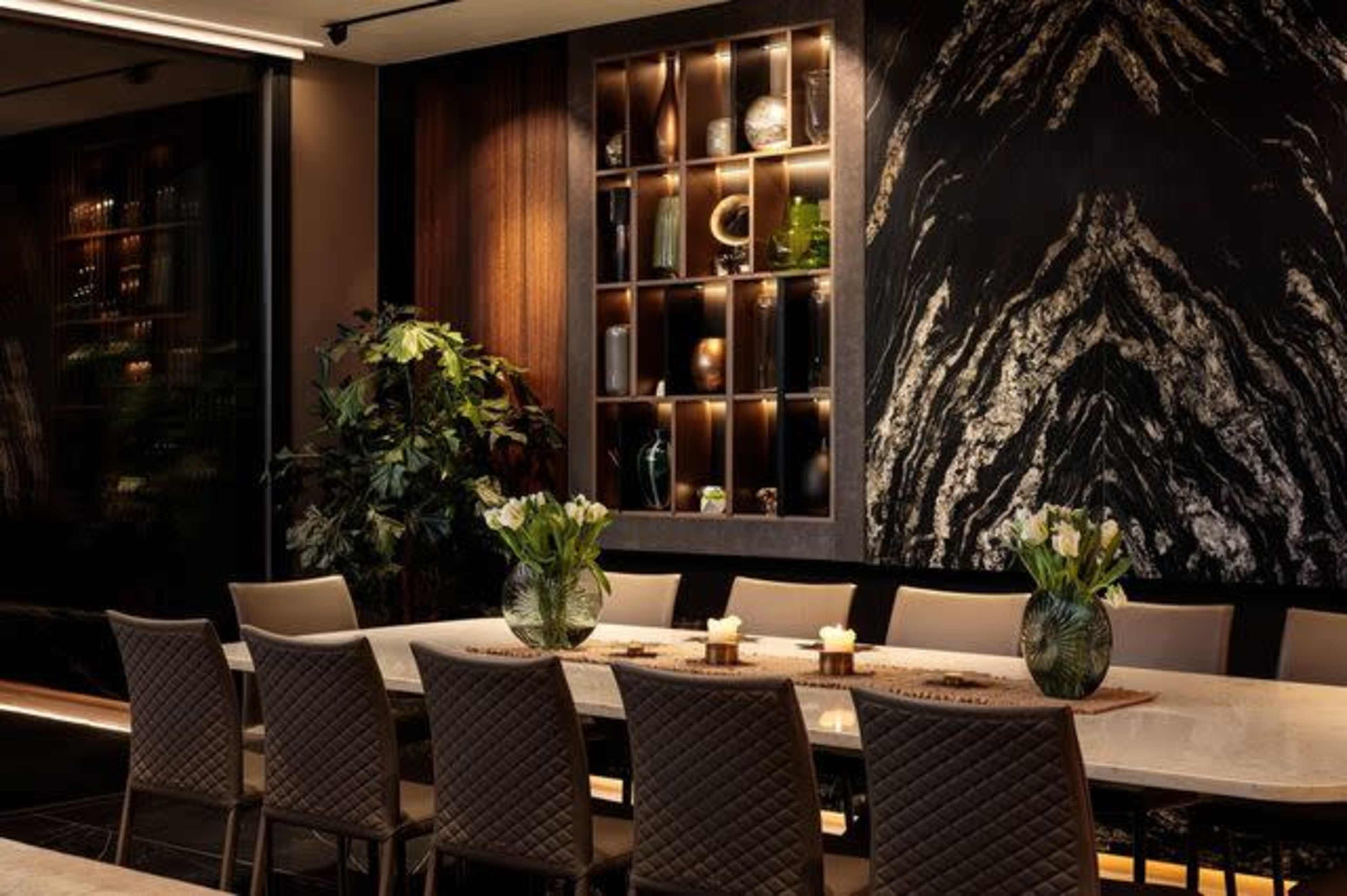 A modern dining room features a long marble table surrounded by upholstered chairs, with a dark stone accent wall and decorative shelves showcasing various objects.
