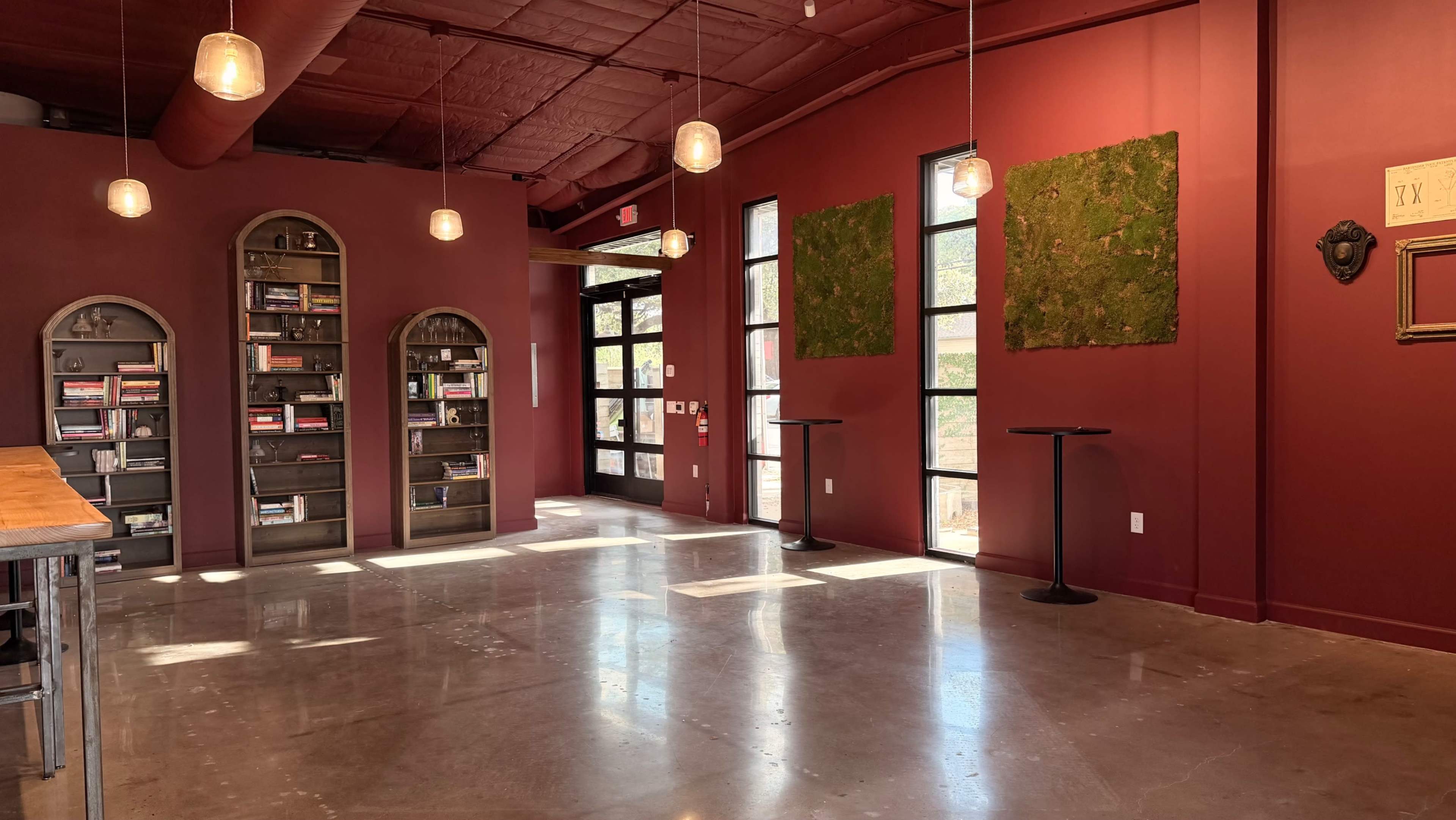 The image shows a spacious interior with a red wall, tall windows, shelves filled with books, and two small round tables.