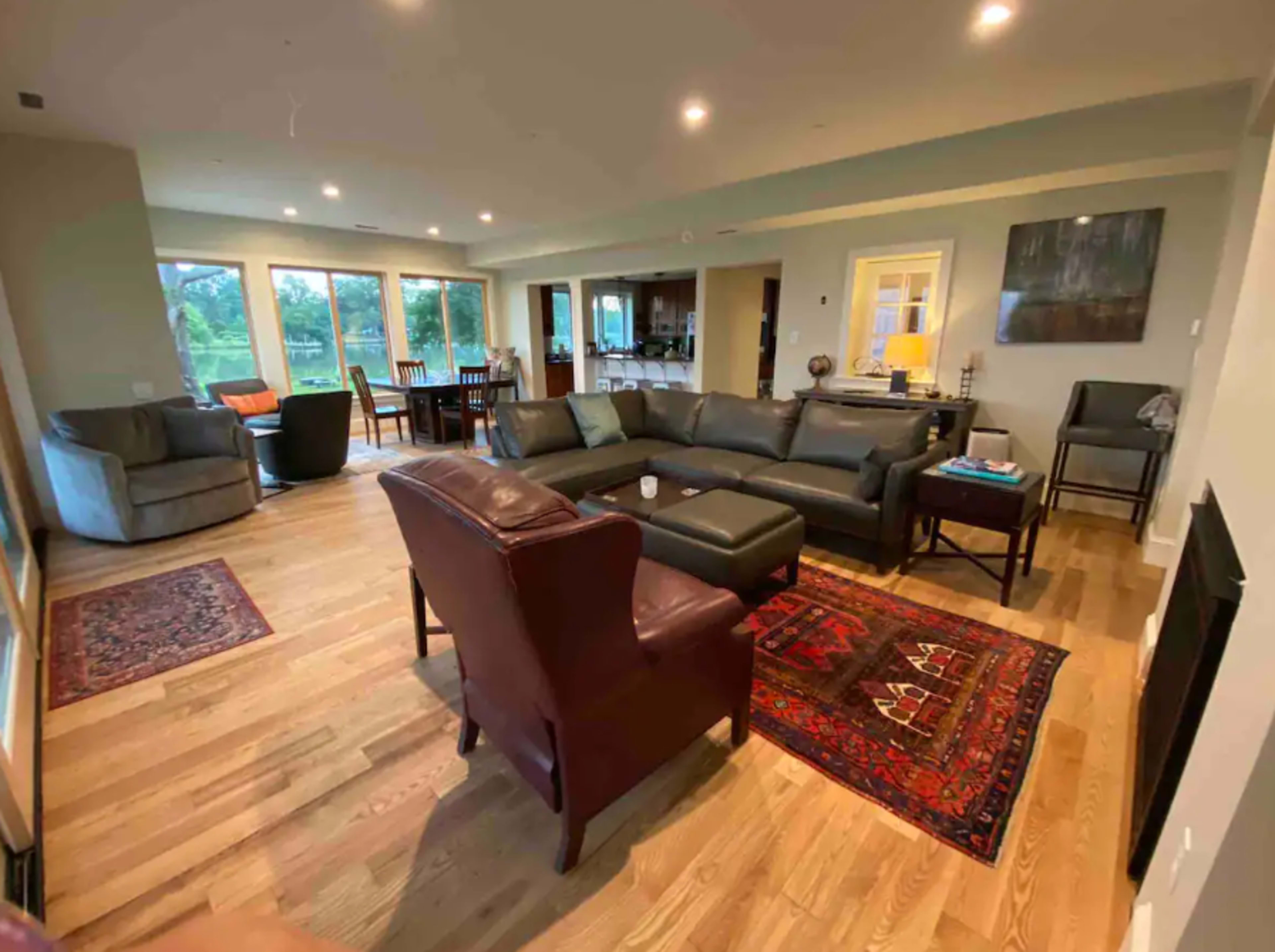 The image shows a spacious living room with large windows, modern furniture, and a combination of hardwood and carpeted flooring.