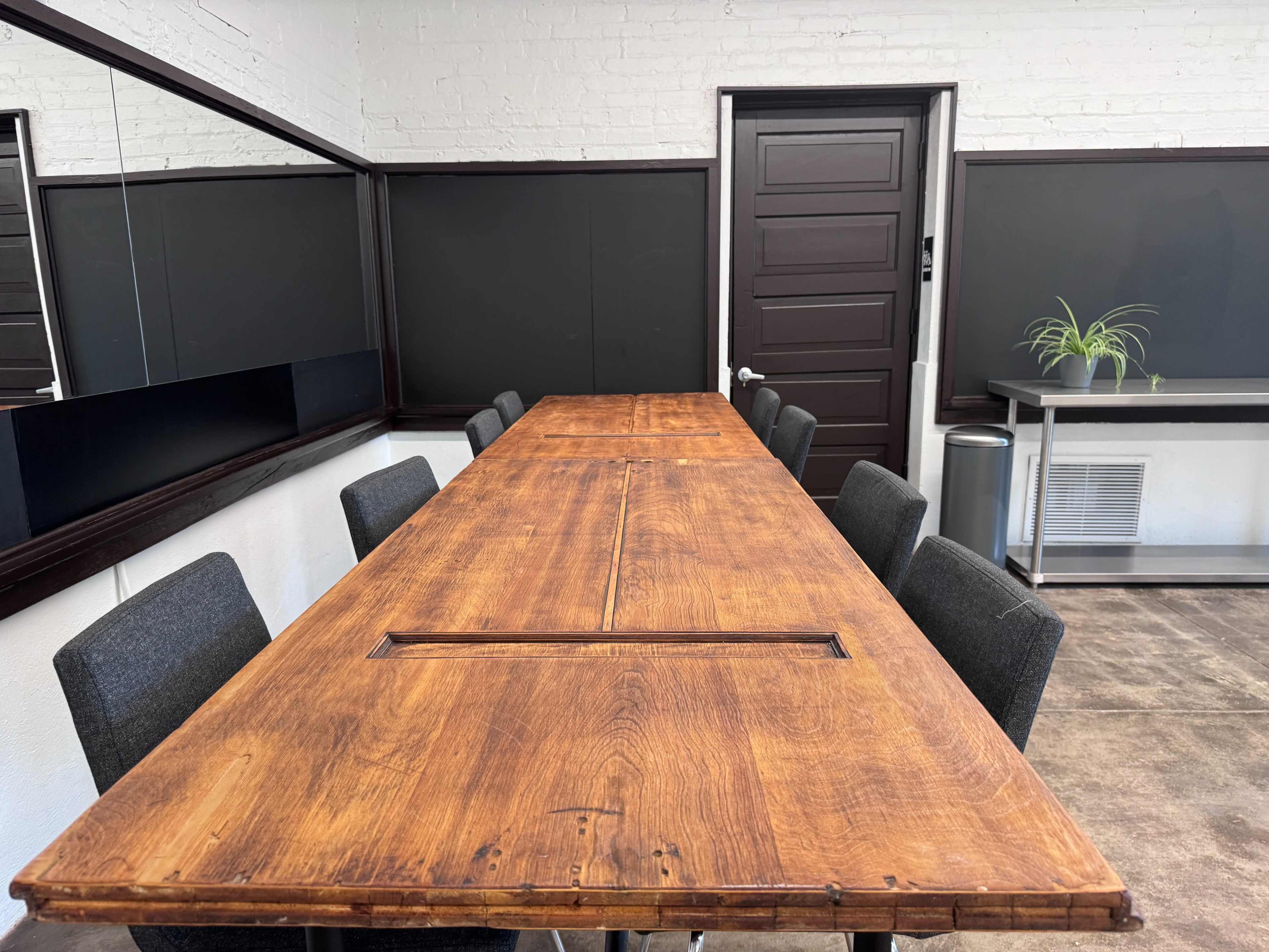 A long wooden conference table is set in a room with dark walls and a large mirror, surrounded by gray chairs.