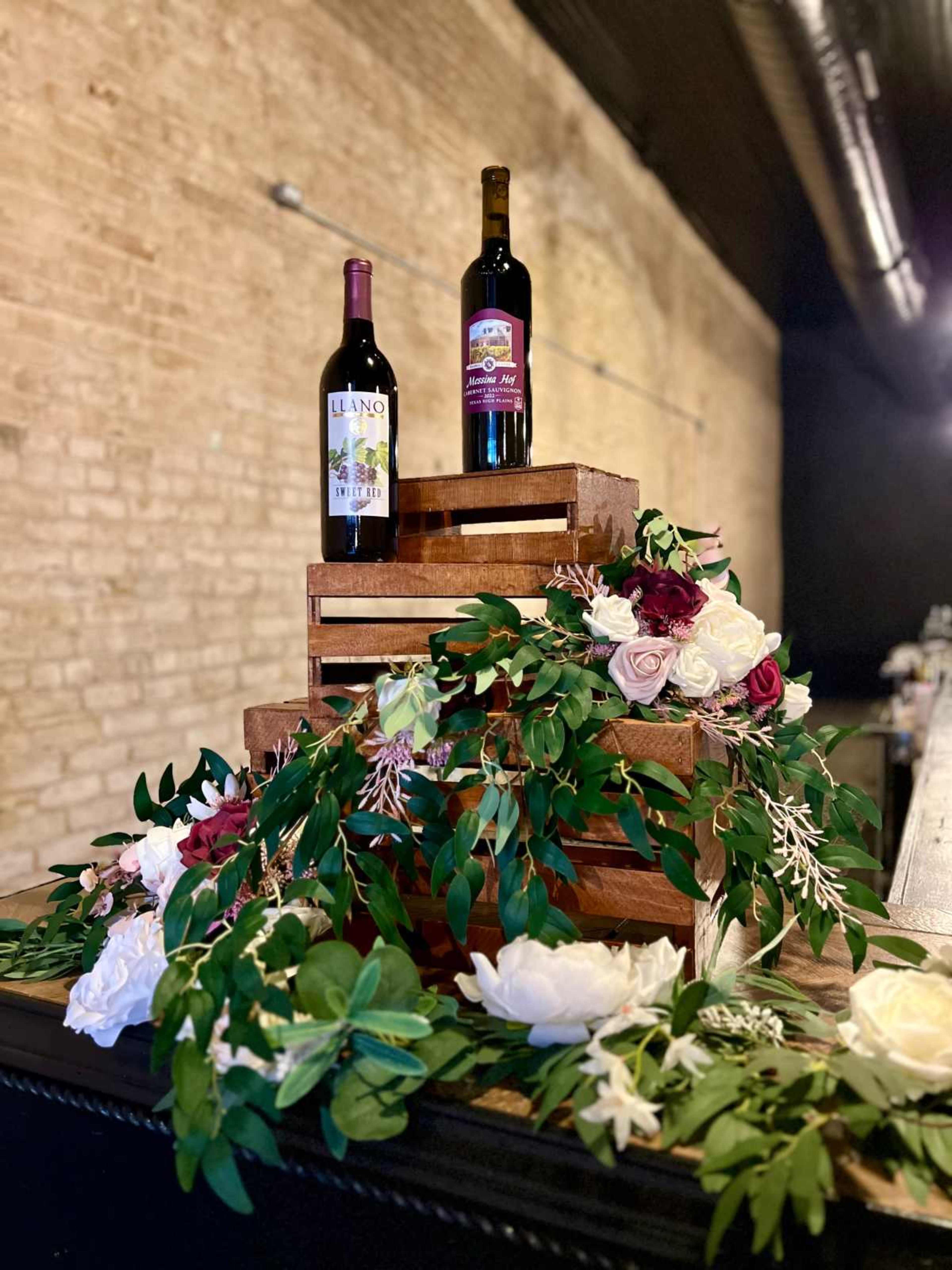 Two bottles of wine are placed on wooden crates, surrounded by floral decorations and greenery.