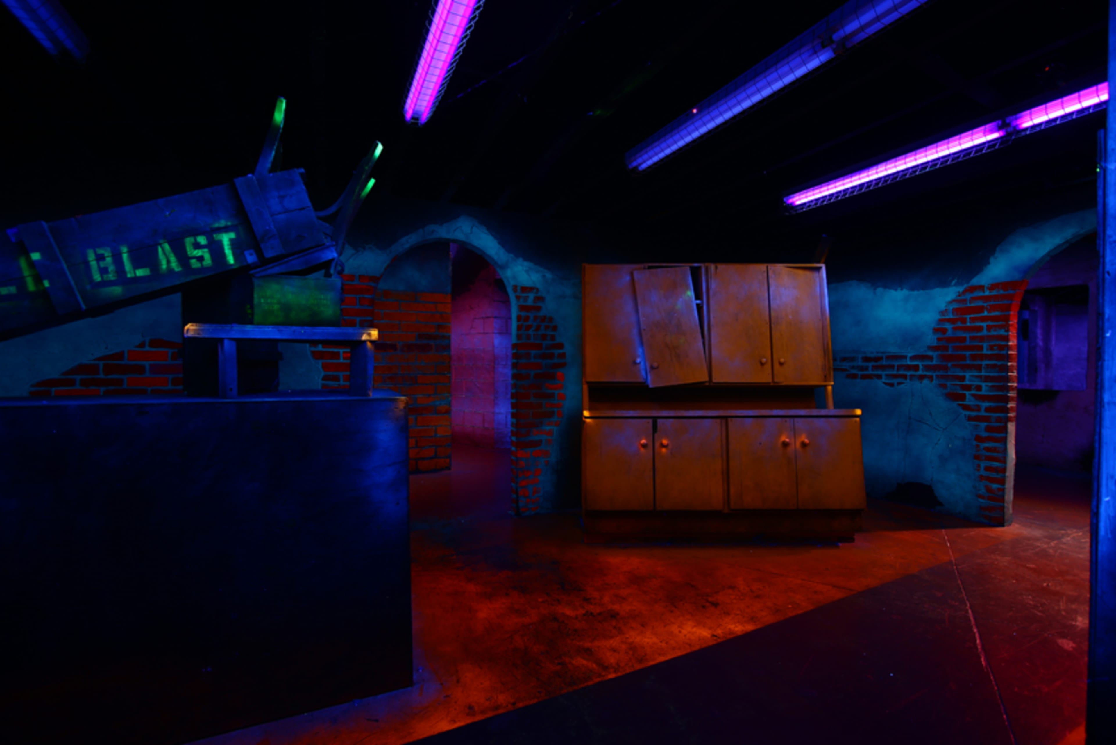 The image shows a dimly lit room with purple and green lighting, featuring a large cabinet and a brick wall in the background.