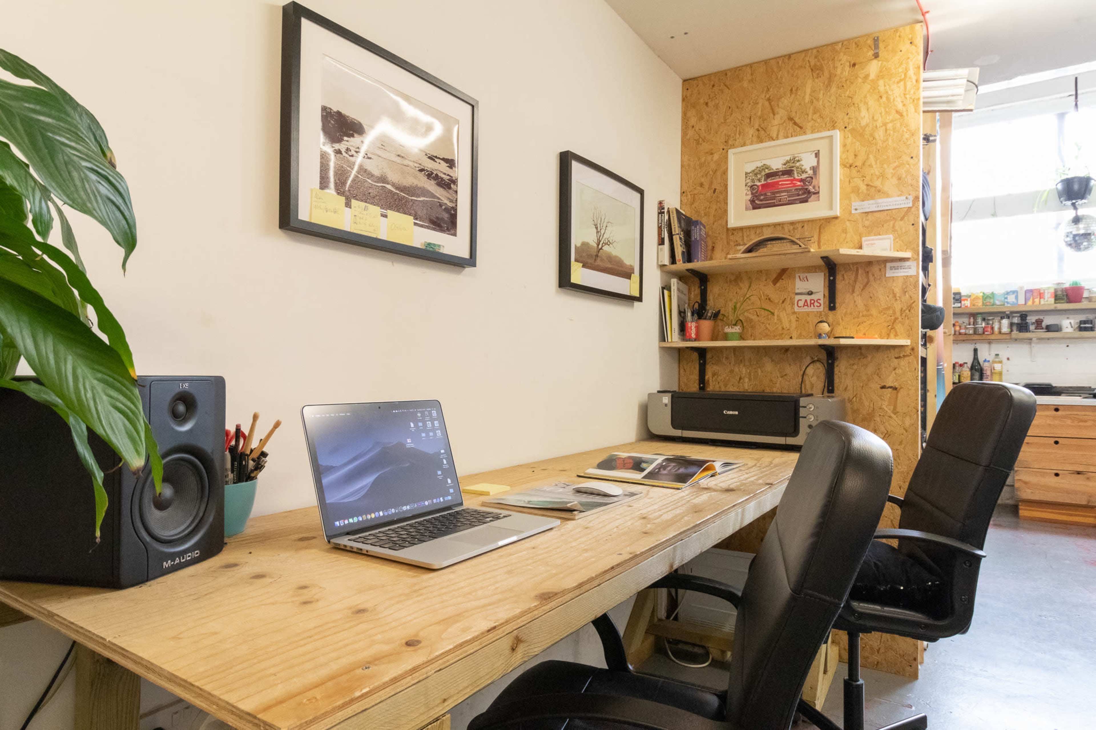 A modern workspace featuring a wooden desk with a laptop, speakers, and two black office chairs against a wall adorned with framed photographs and shelves.