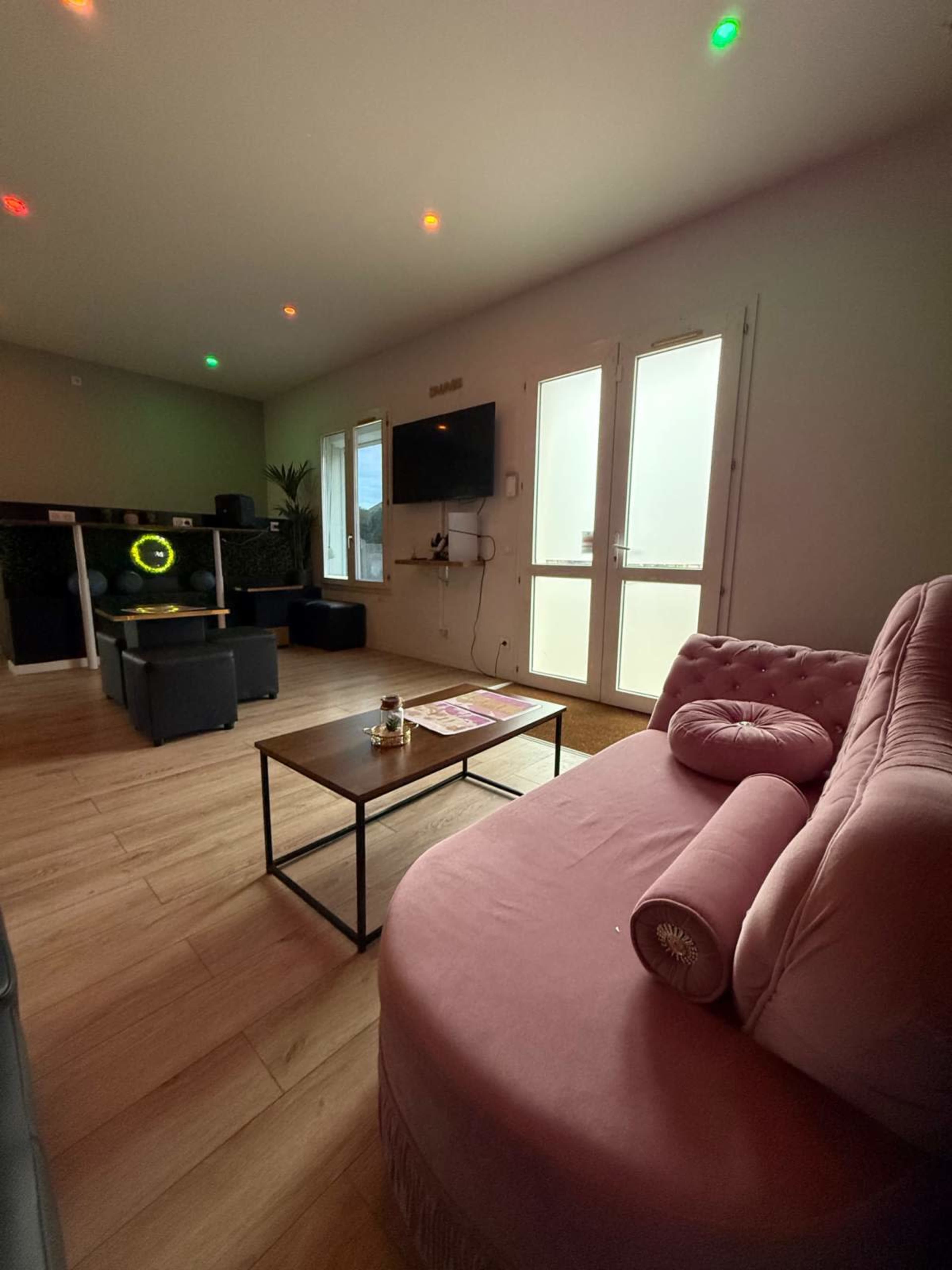 The room features a pink couch, a wooden coffee table, and a television mounted on the wall with large windows allowing natural light.