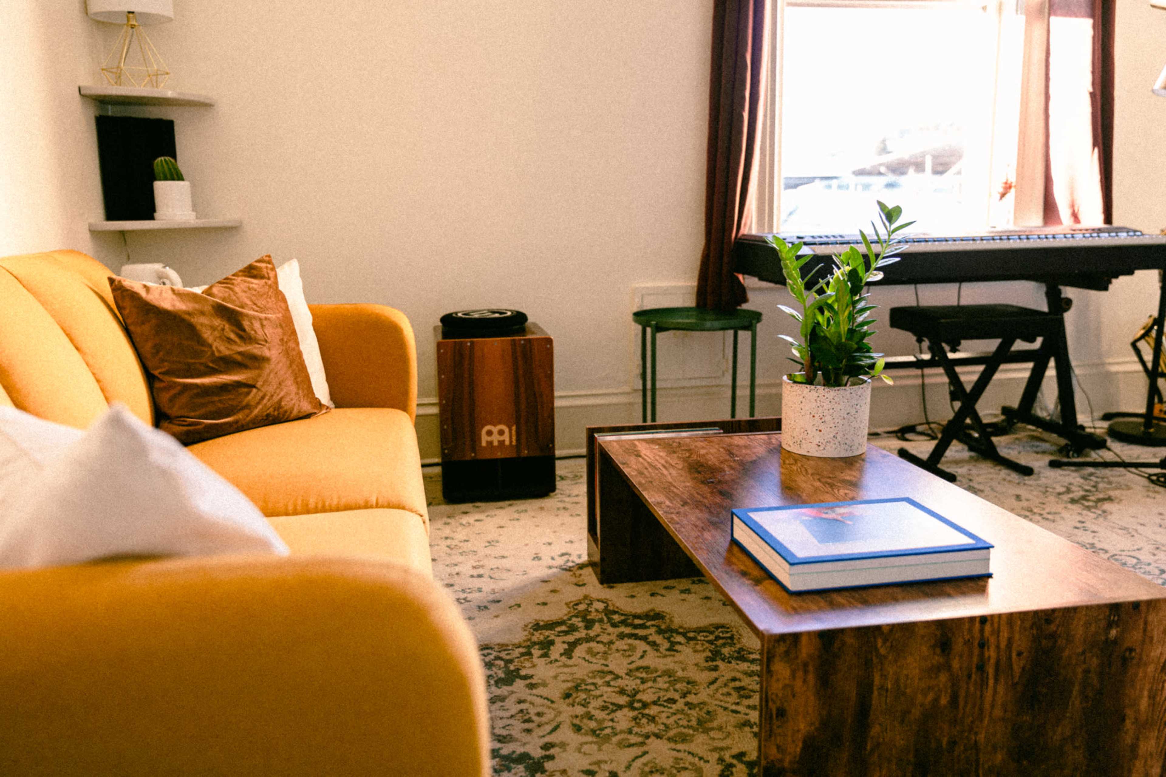 A cozy living room features a yellow sofa, a wooden coffee table with a book, a potted plant, and a keyboard setup near a window.