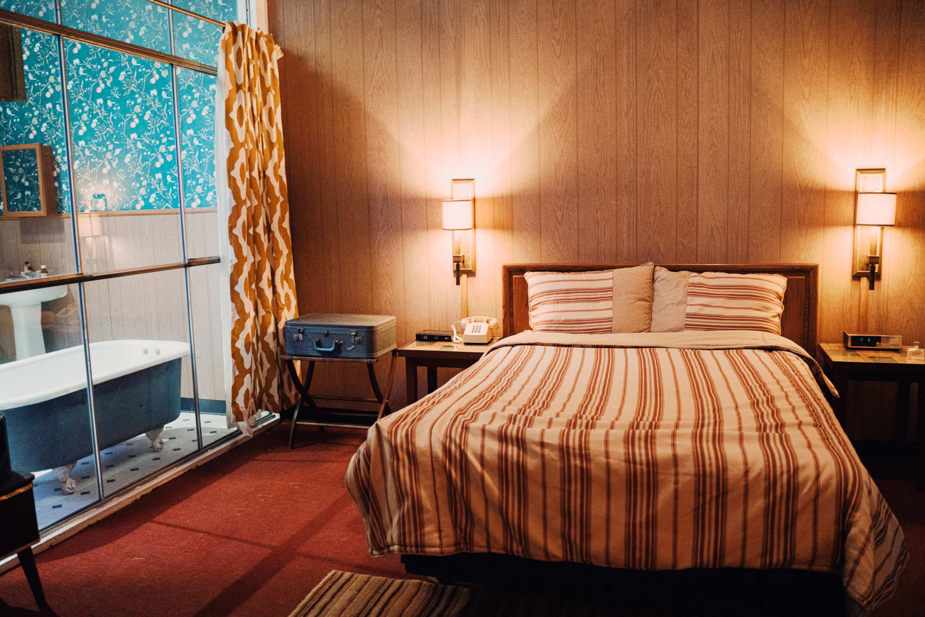 A cozy bedroom with a striped bedspread, two bedside lamps, a vintage-style telephone, and a visible bathroom area featuring a freestanding bathtub.