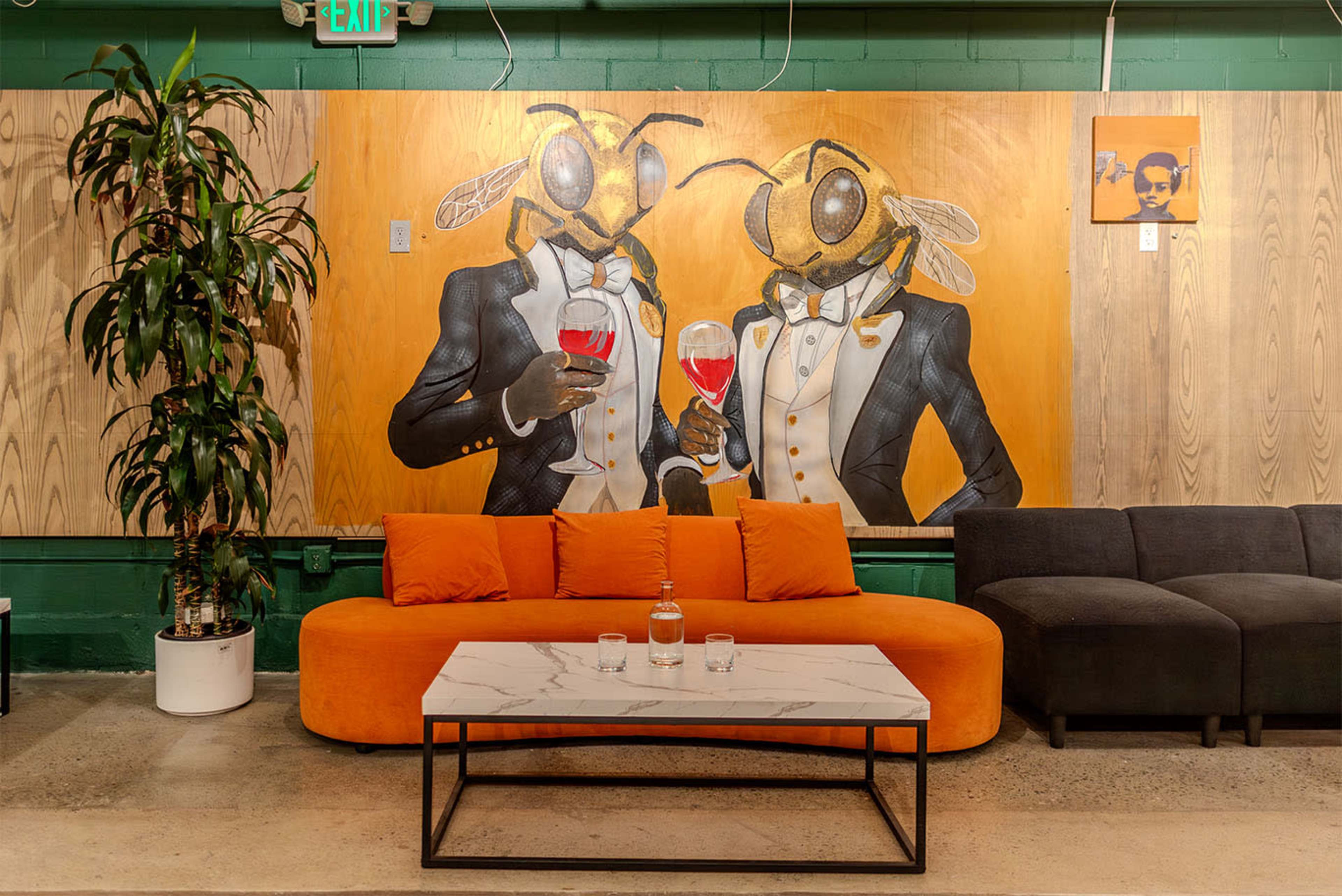 The image depicts a colorful mural of two anthropomorphic bees in tuxedos holding glasses of red wine, with a cozy seating area featuring a marble coffee table and an orange couch.