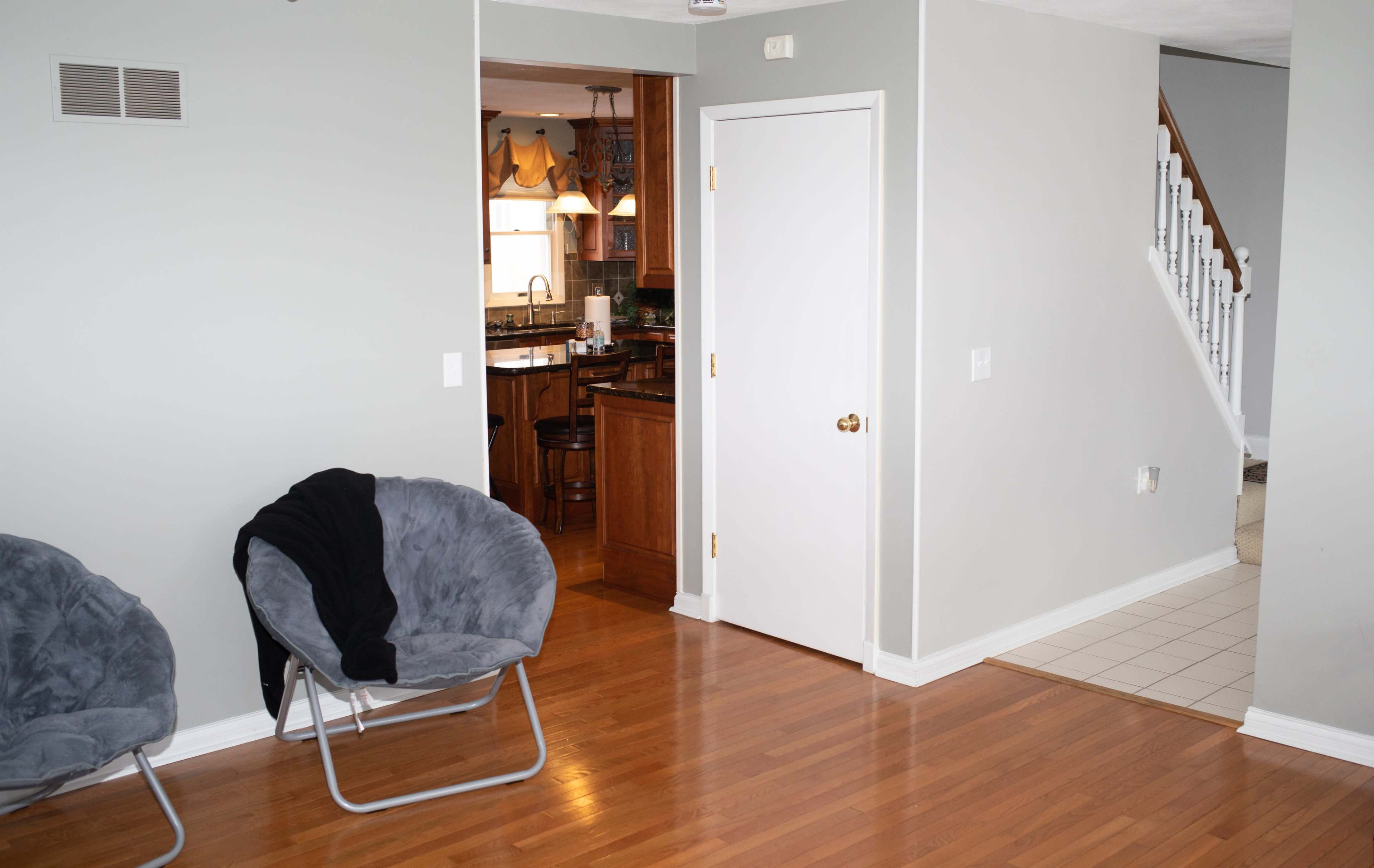 The image shows a cozy living space featuring a gray chair, an open doorway leading to a kitchen, and a staircase in the background.