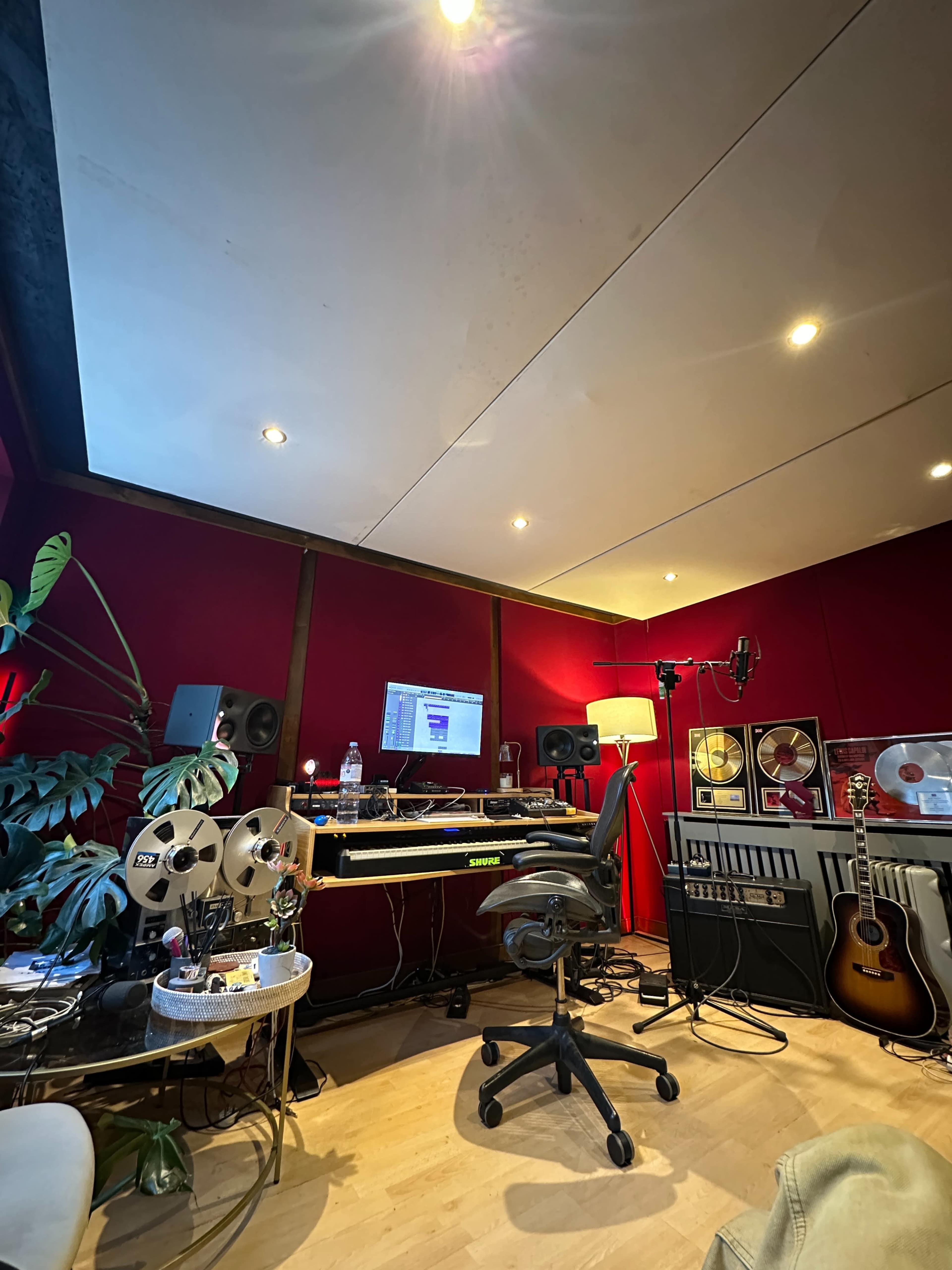 The image shows a music recording studio featuring a desk with recording equipment, a computer screen, various audio monitors, a guitar, and red walls.
