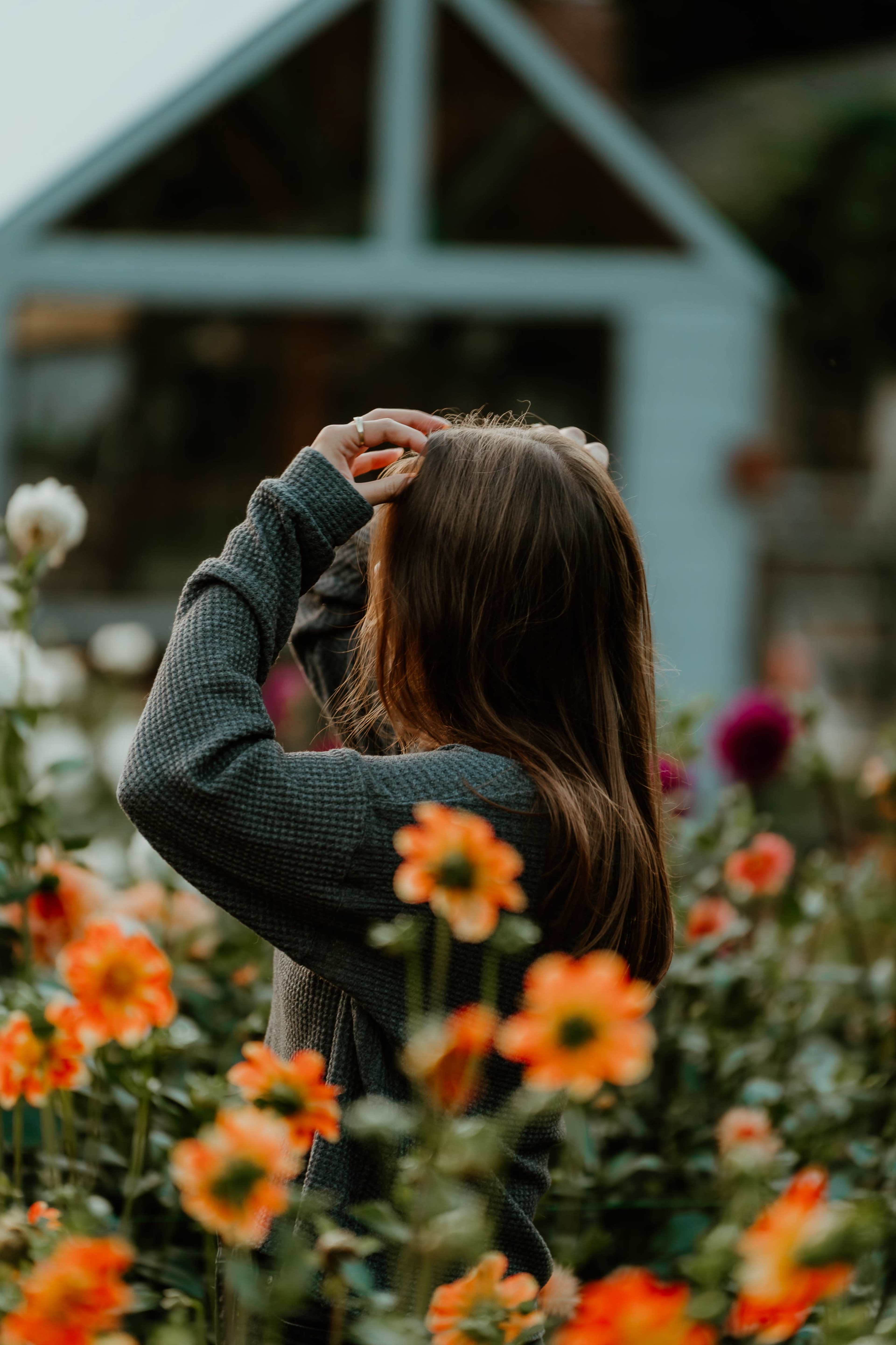A woman stands in a garden surrounded by colorful flowers, brushing her hair back as she gazes away from the camera.