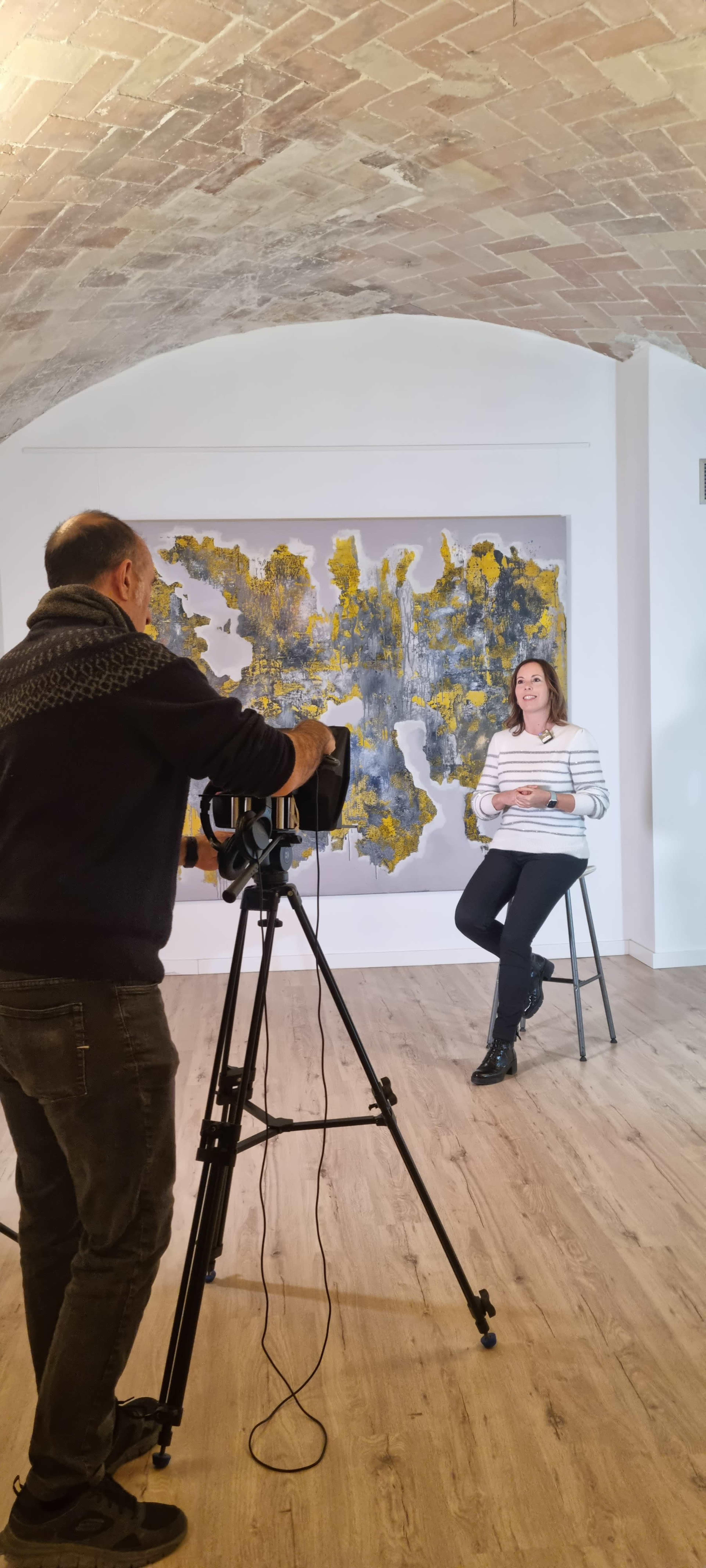 A woman sits on a stool in front of a large abstract painting while a man operates a camera on a tripod directed at her.