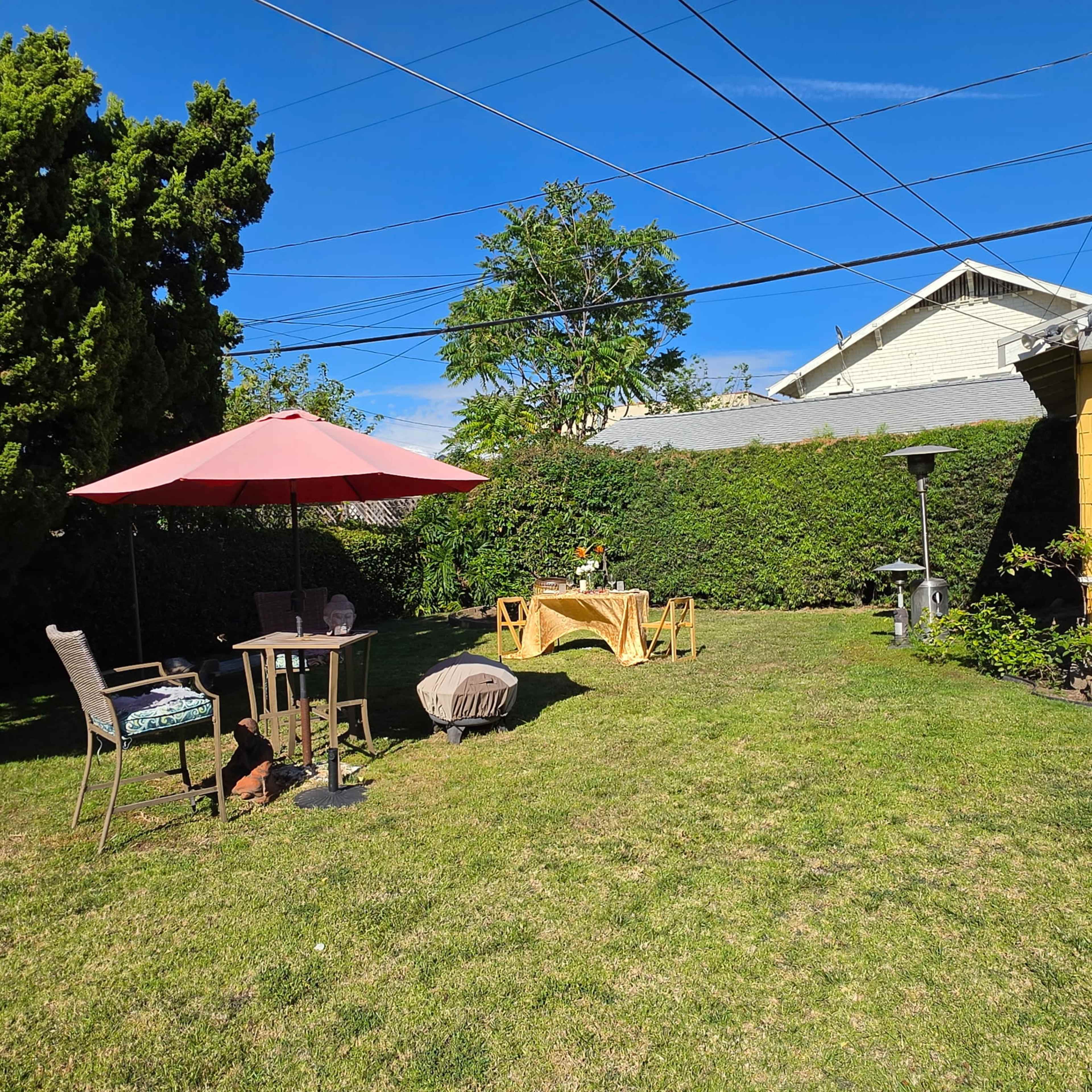The image shows a backyard with a red umbrella over a small table and chairs, a decorative table set up, and greenery surrounding the area.