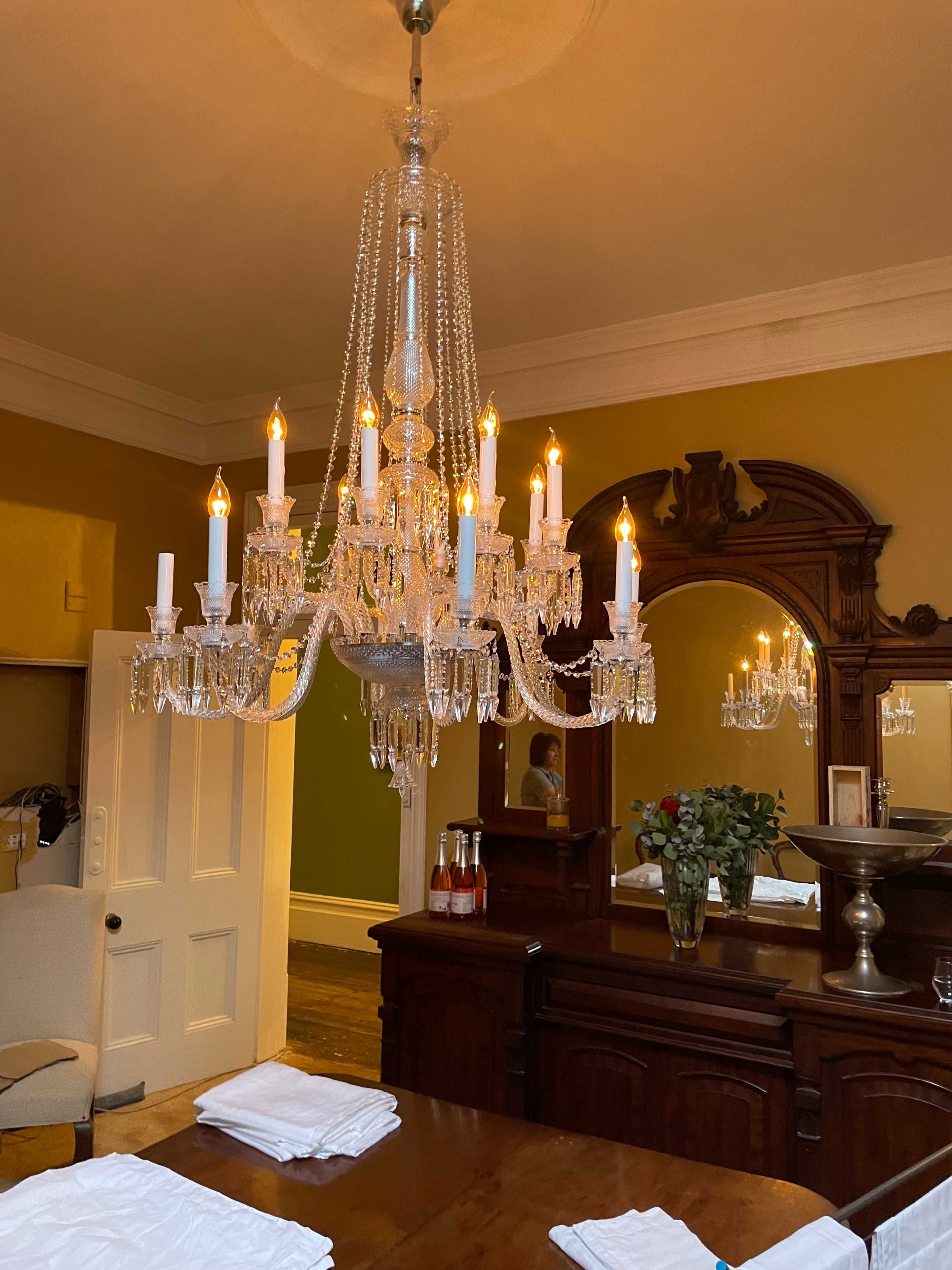 A crystal chandelier hangs from the ceiling above a wooden dining table in a room featuring a large mirror and an ornate wooden sideboard.