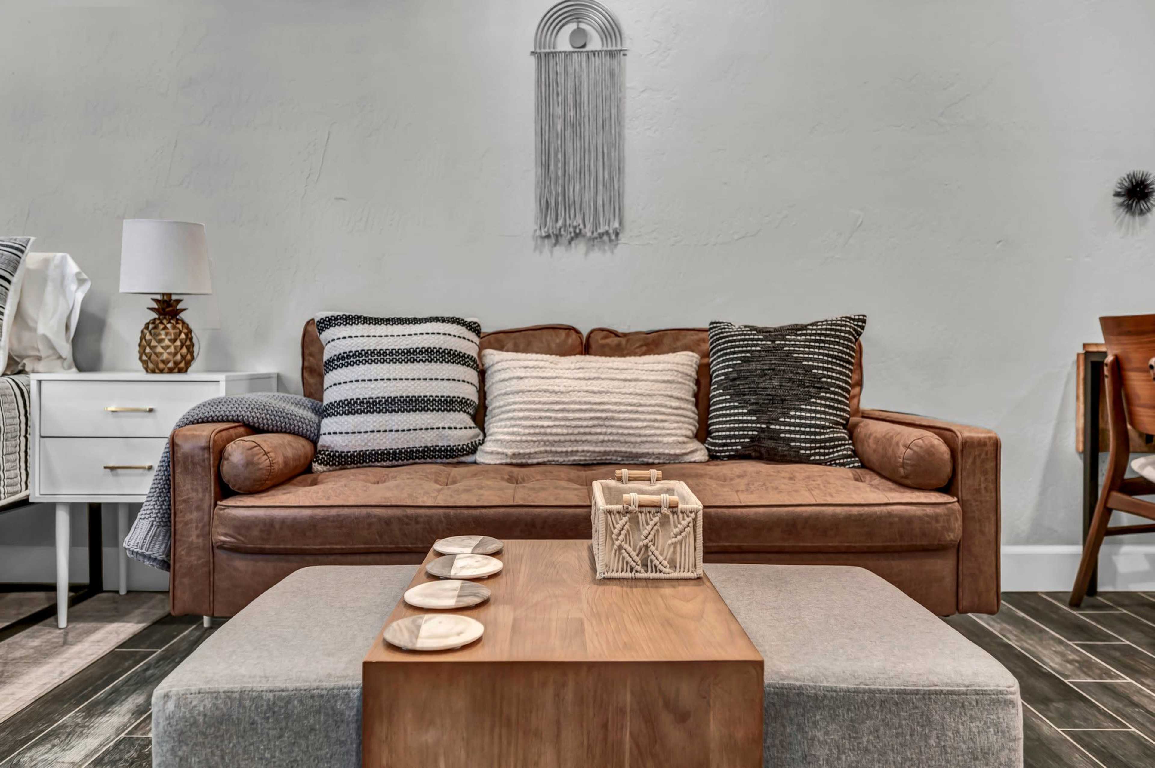 A brown leather sofa with patterned cushions is positioned in a living room, facing a wooden coffee table and surrounded by neutral-toned decor.