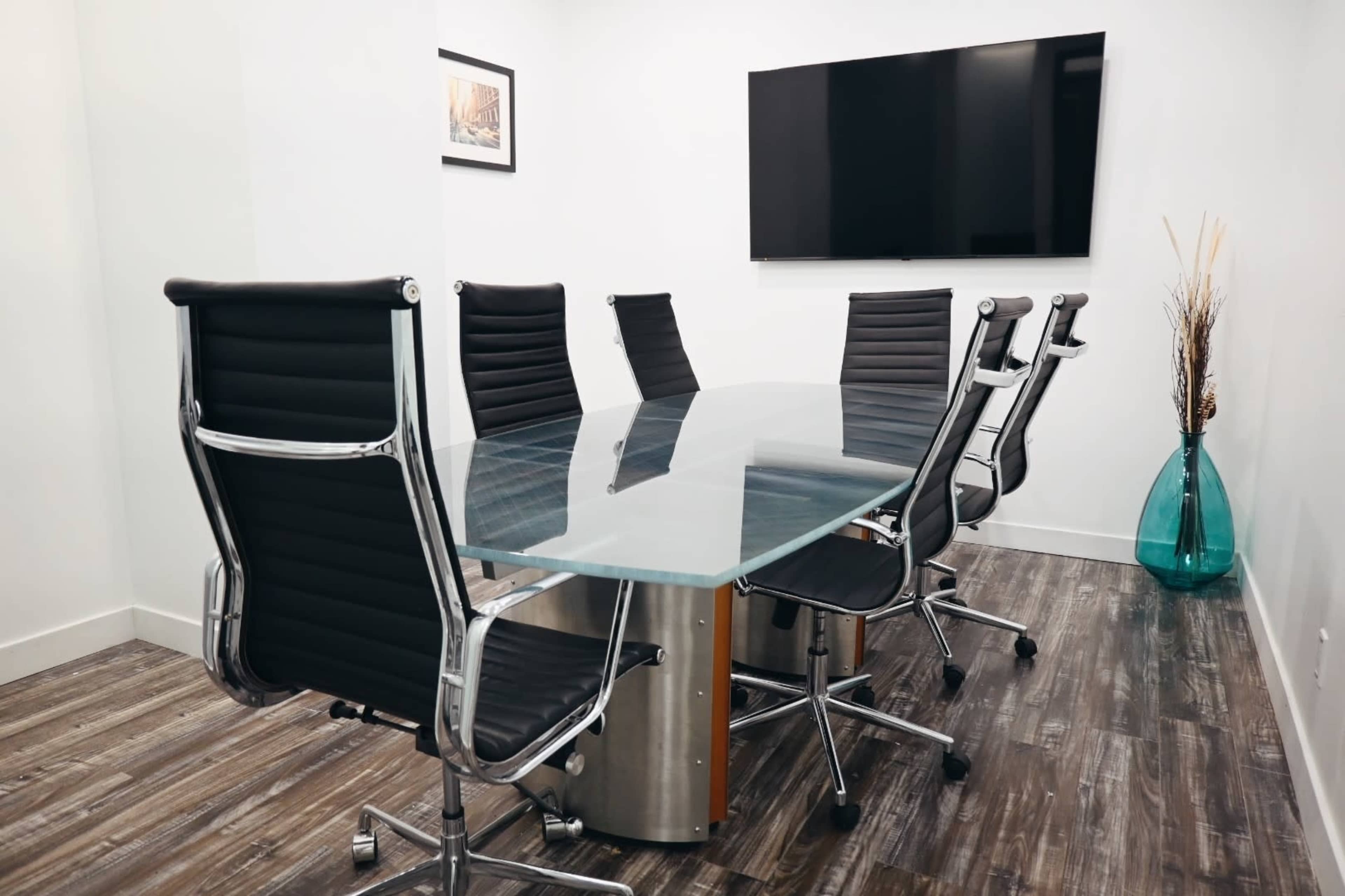 A modern conference room features a glass table surrounded by black leather swivel chairs, with a flat-screen TV mounted on the wall and a decorative vase in the corner.