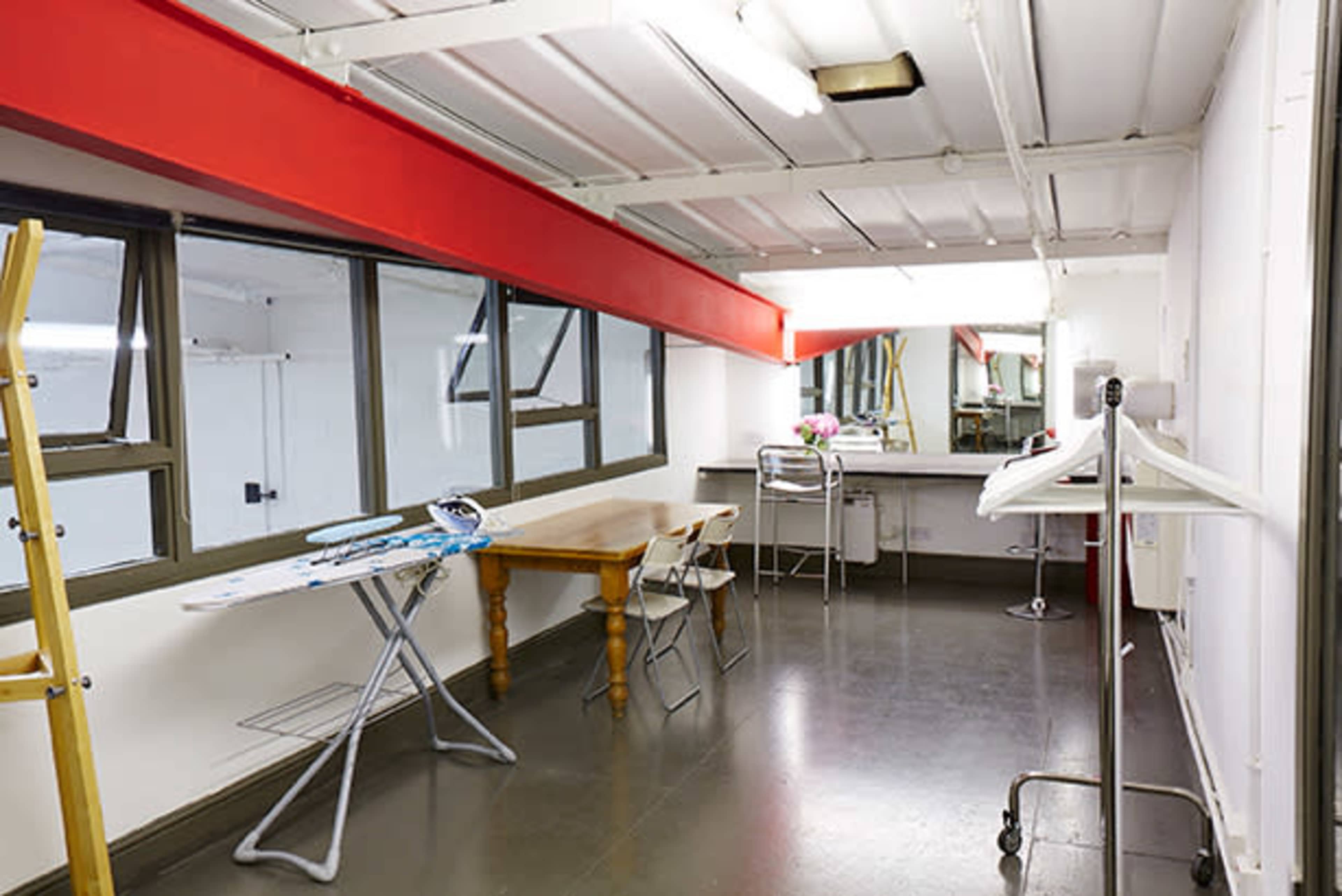 A bright, minimalist studio space featuring a wooden table, an ironing board, and large windows with a red accent wall.