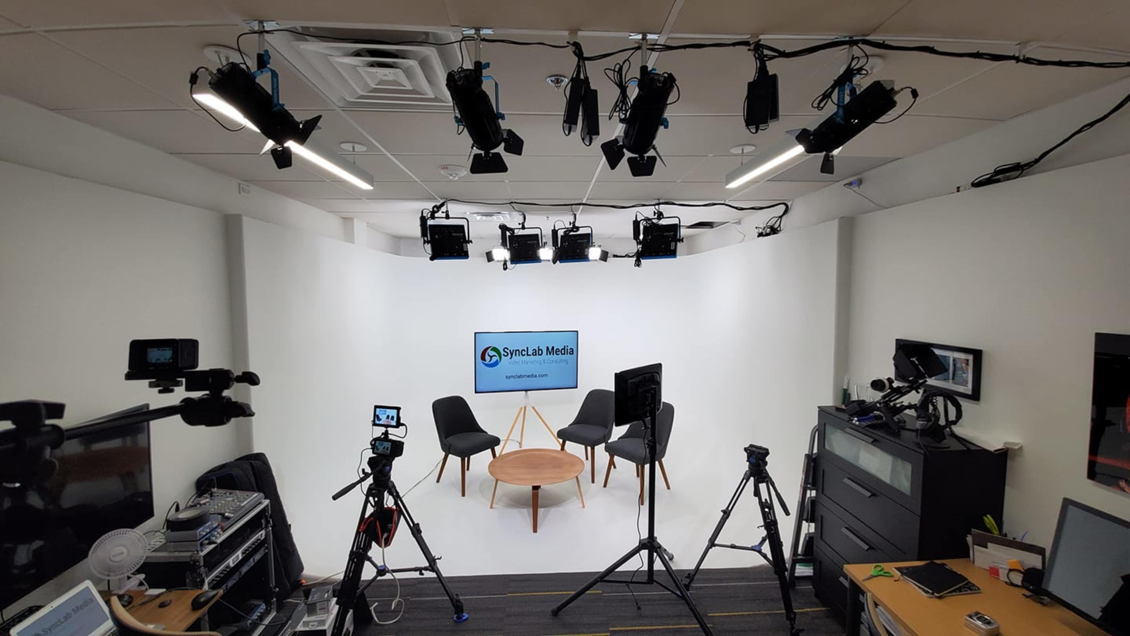 A media studio setup with two chairs, a central table, multiple cameras, and studio lighting against a white backdrop featuring a logo for "Syndicate Media."