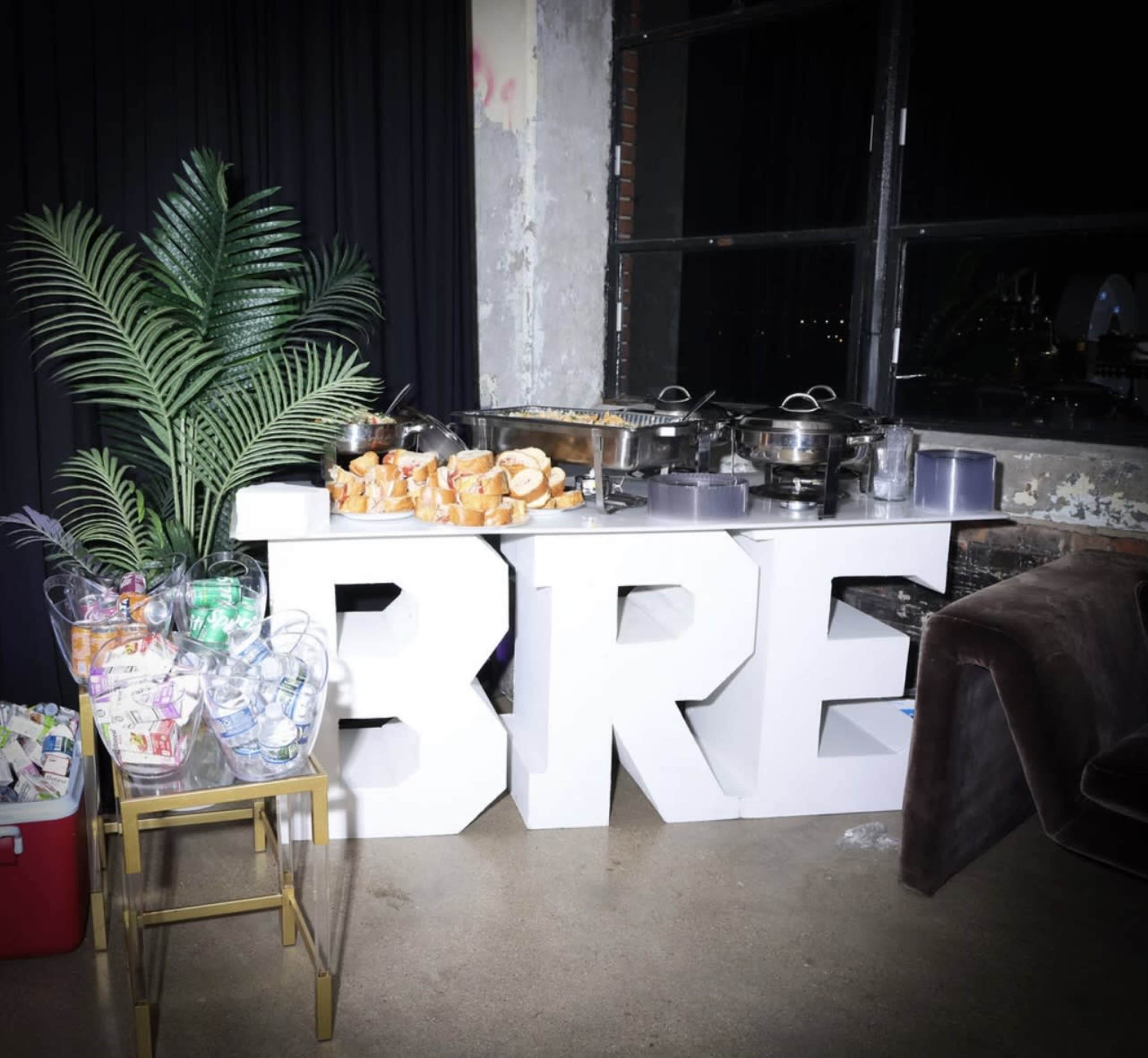 A food display featuring large letters spelling "BRE" is set up beside a decorative plant and various catering trays.