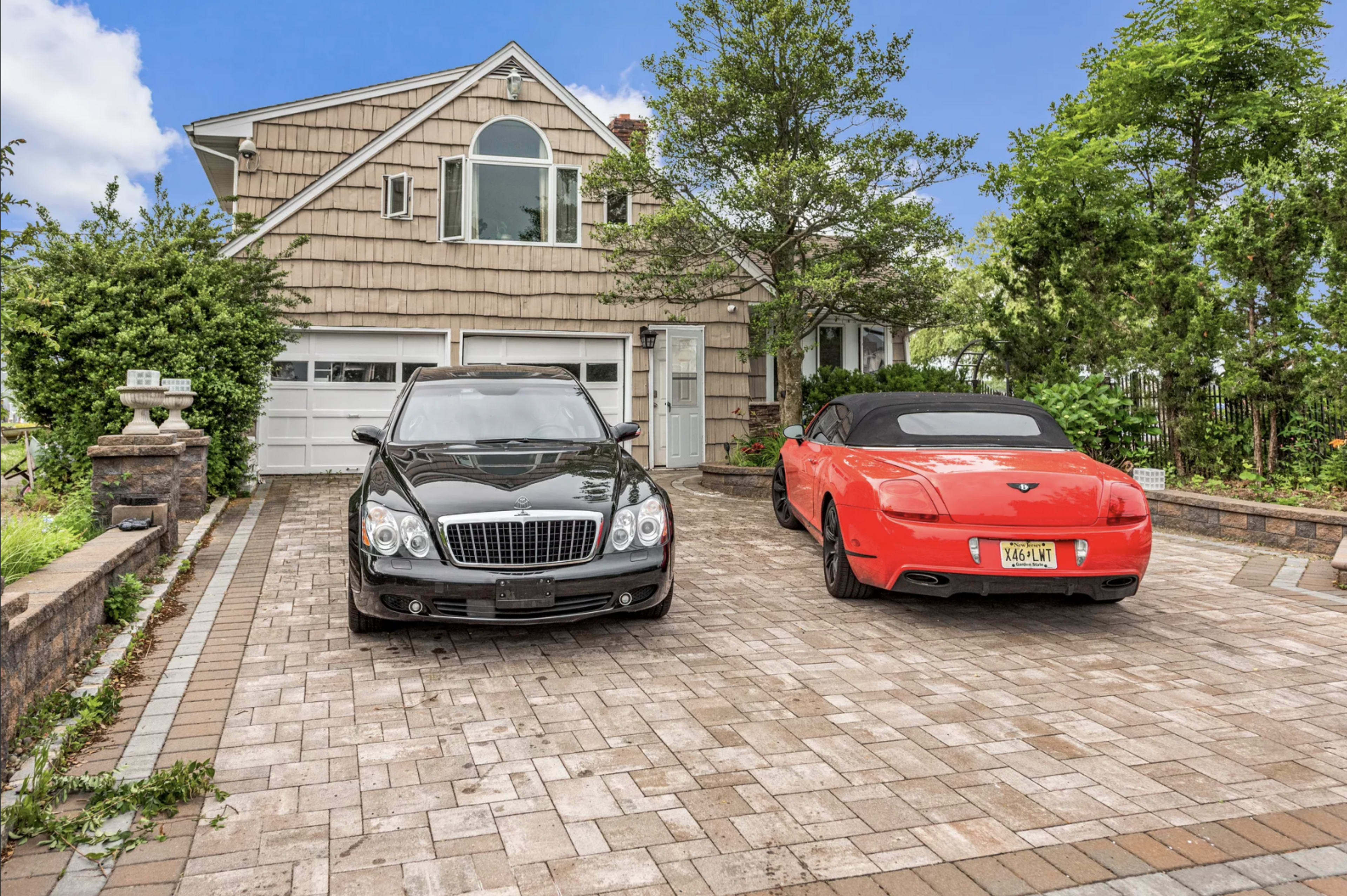 A black luxury sedan and a red convertible are parked in front of a house with a shingle exterior and a paved driveway.