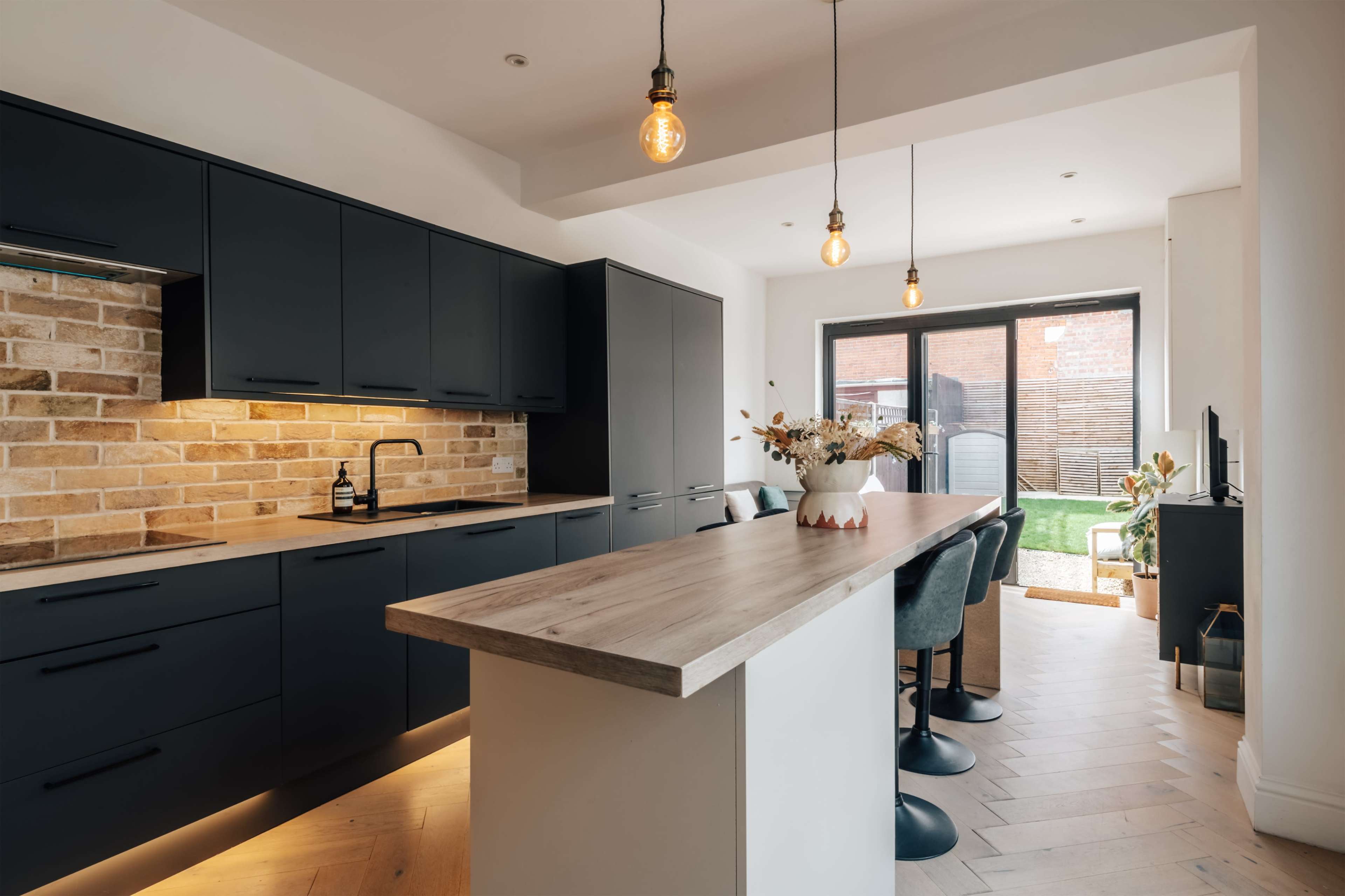 The image shows a modern kitchen with dark cabinetry, a light wood island, pendant lights, and a sliding door leading to a backyard.