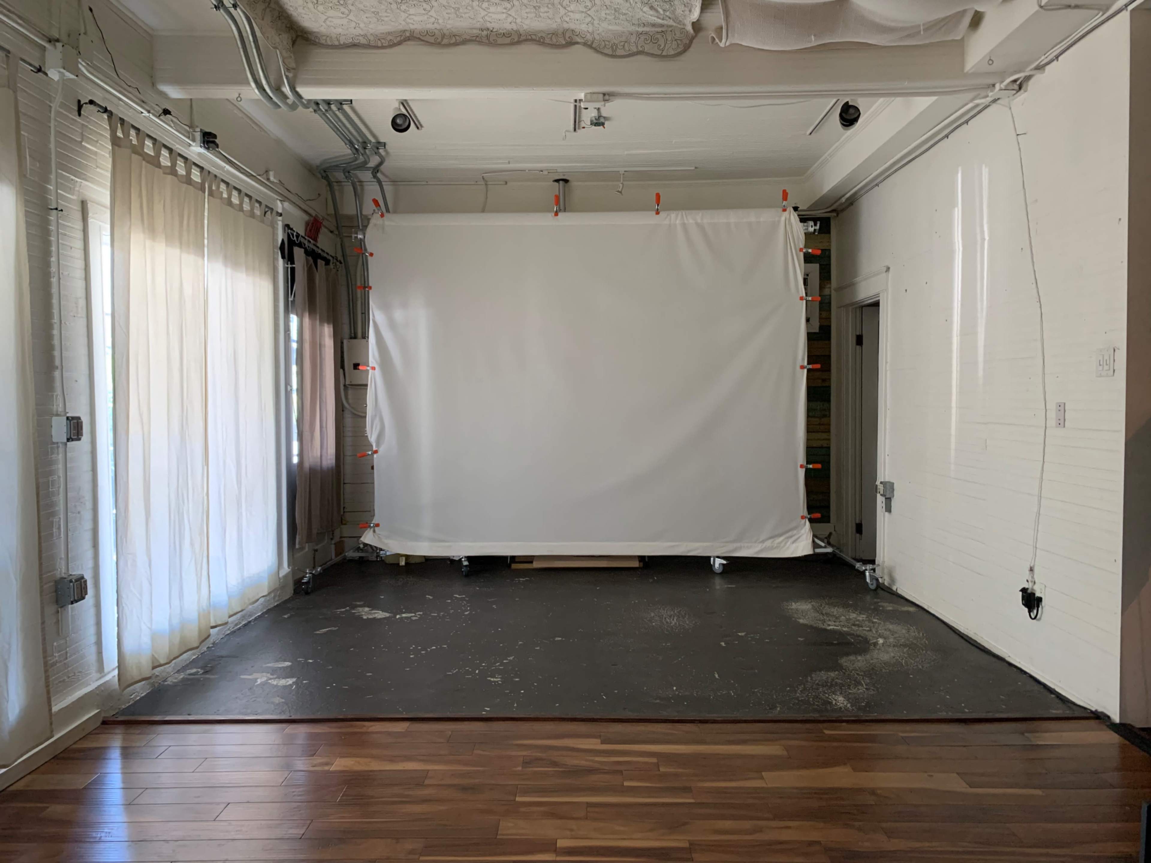 The image shows an empty room with a white backdrop set up on wheels, surrounded by natural light from large windows and wooden flooring.