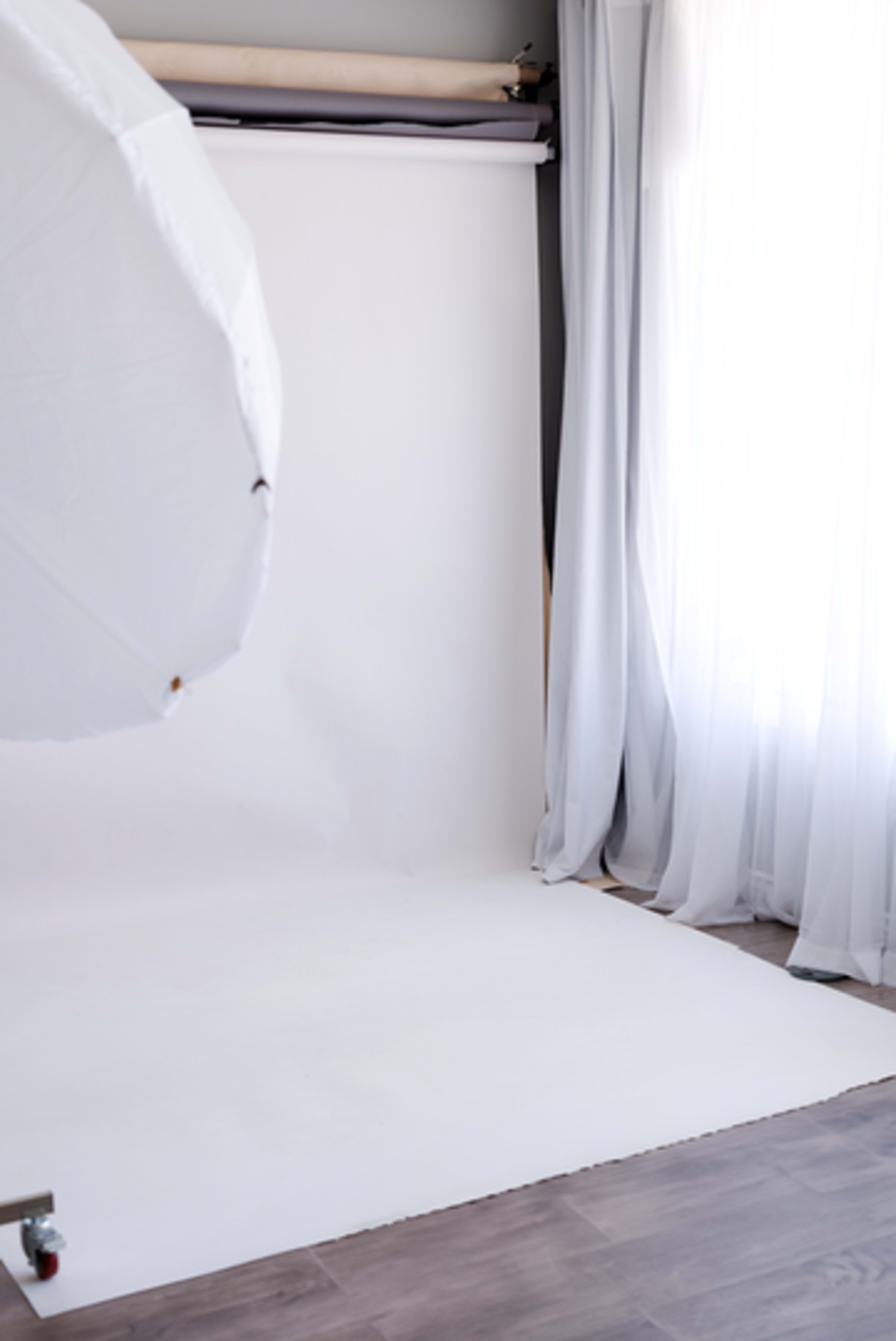 The image shows a photography studio with a white backdrop, natural light coming through sheer curtains, and a softbox partially visible in the foreground.