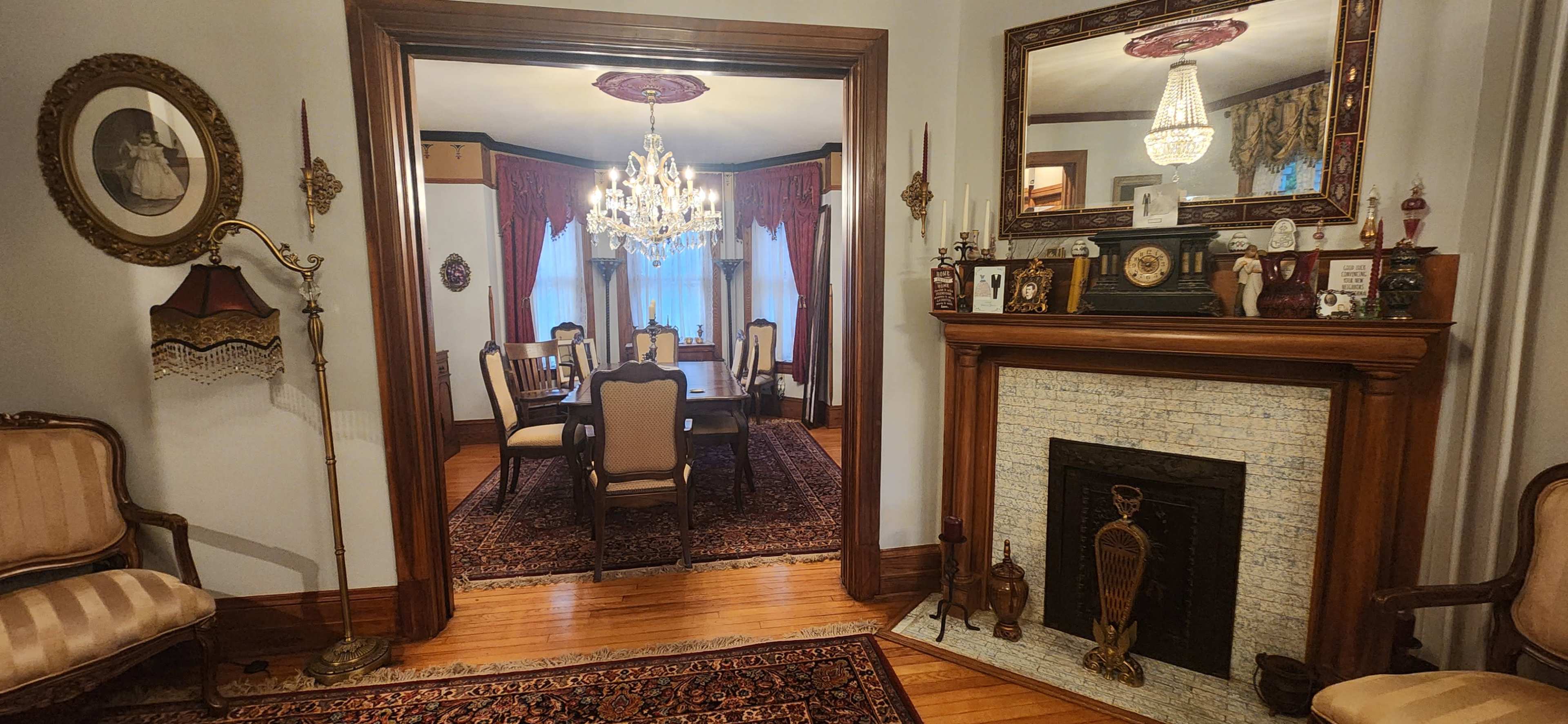 The image shows a view from a living room into a formal dining room, featuring ornate furnishings, a chandelier, and a fireplace with decorative elements.