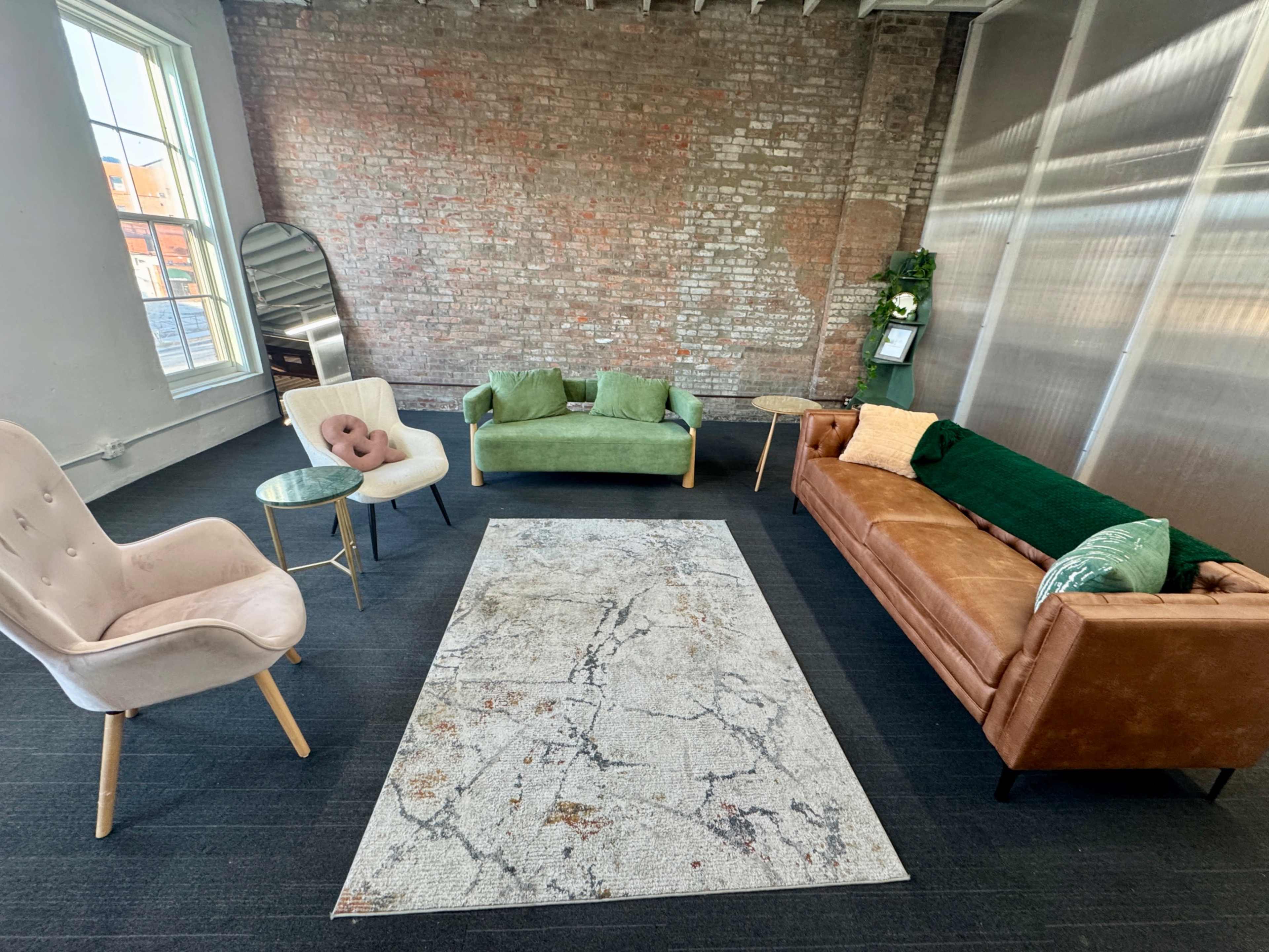 The image shows a spacious, minimalist living area with a brown leather sofa, a green couch, a pink chair, two small side tables, and a large patterned rug on a dark carpeted floor.