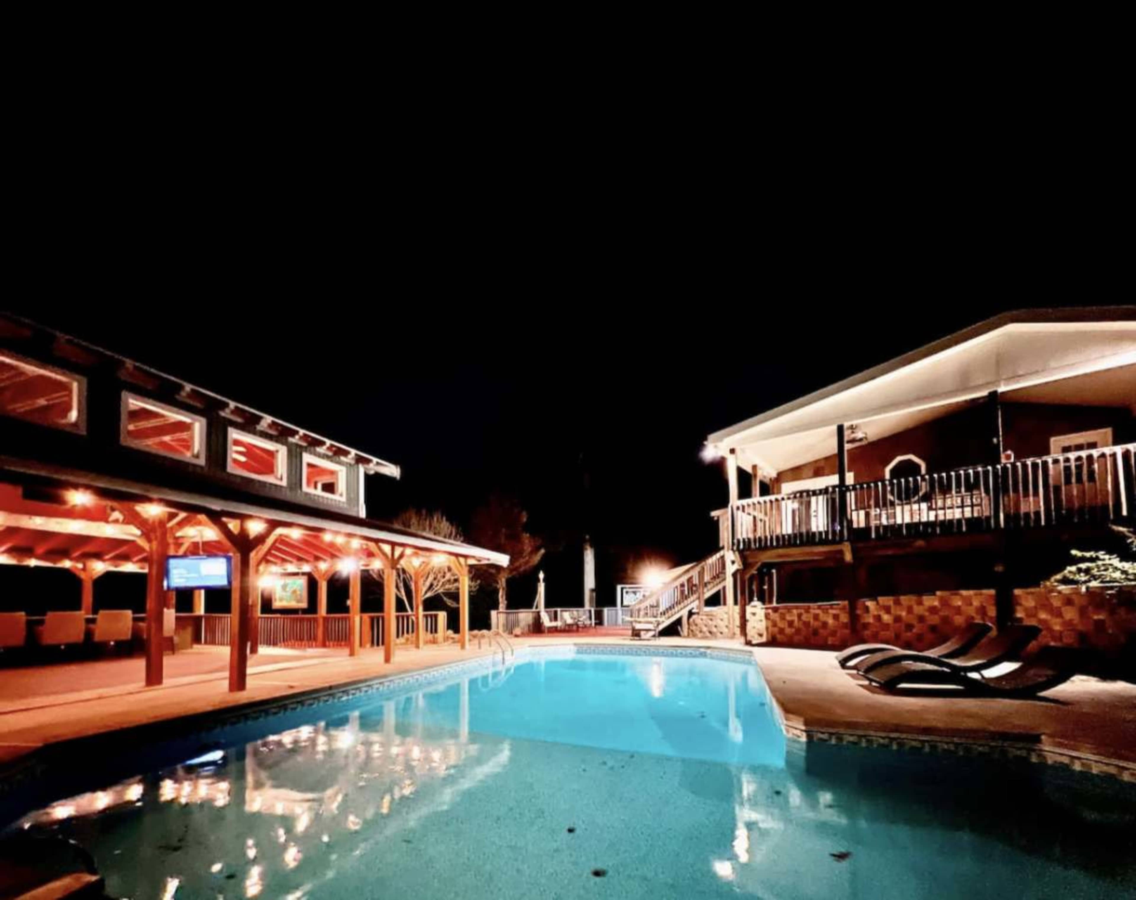 The image shows a well-lit swimming pool surrounded by a deck and two buildings, with a festive nighttime atmosphere.